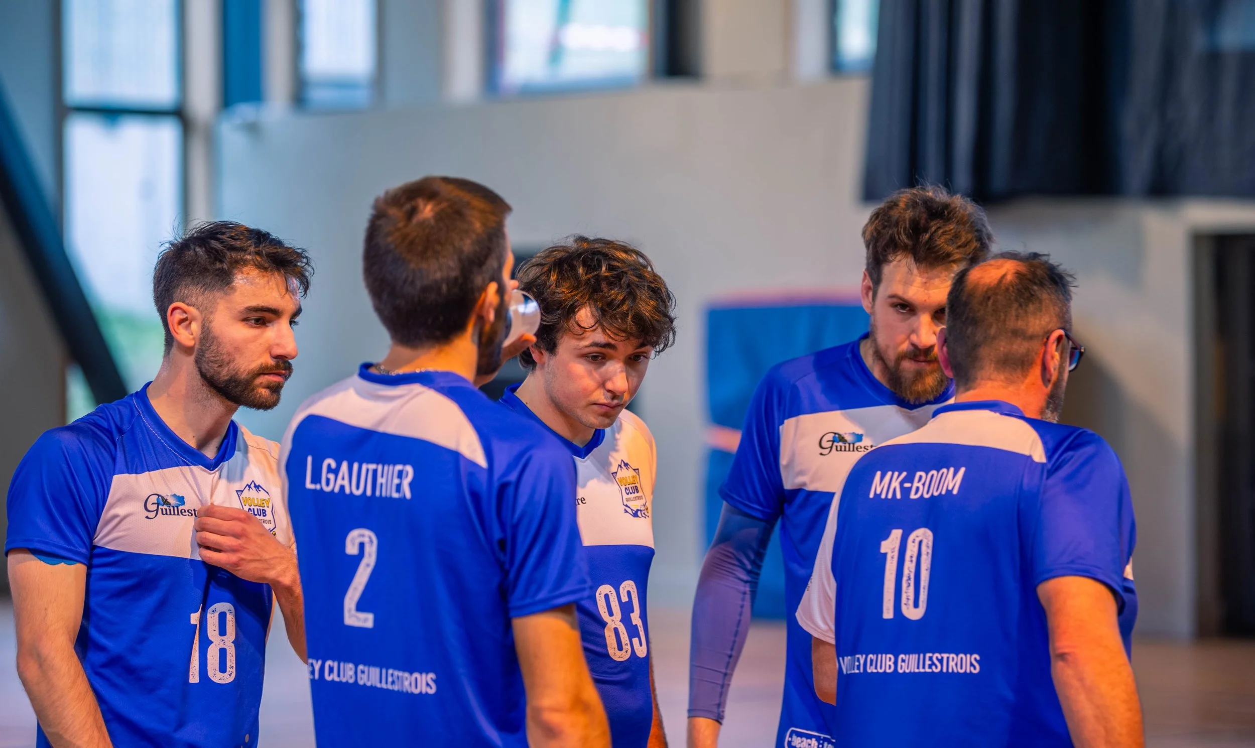 Cinq joueurs de volley-ball en réunion, portant des maillots bleus avec des numéros, dans une salle intérieure.