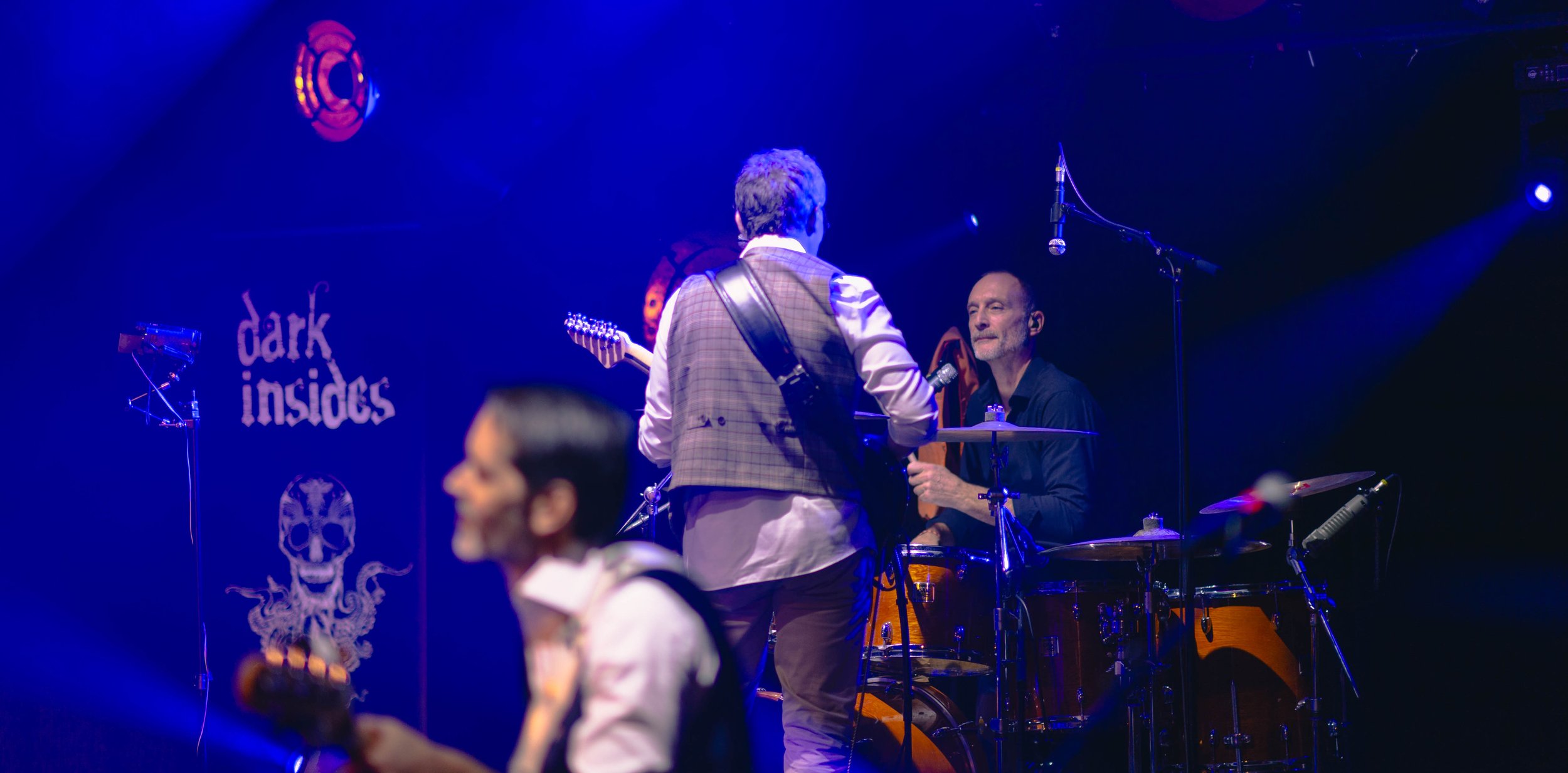 Trois musiciens sur scène en train de jouer, avec un fond illustrant un logo 'dark insides' et un crâne. La scène est éclairée de lumière bleue.