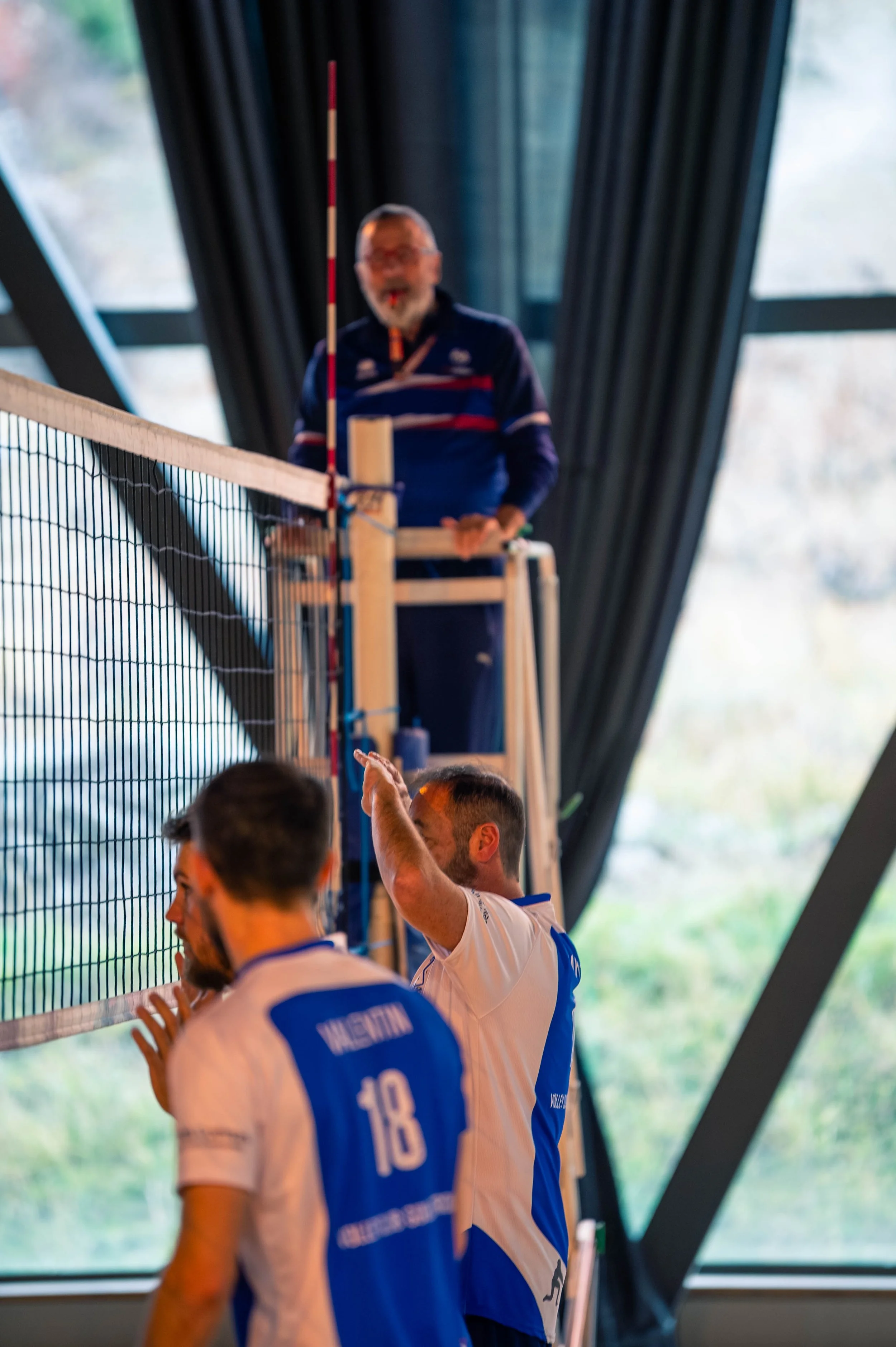 Deux joueurs de volley-ball en train de discuter, avec un arbitre suspendu au-dessus d'eux dans une salle intérieure avec de grandes fenêtres.