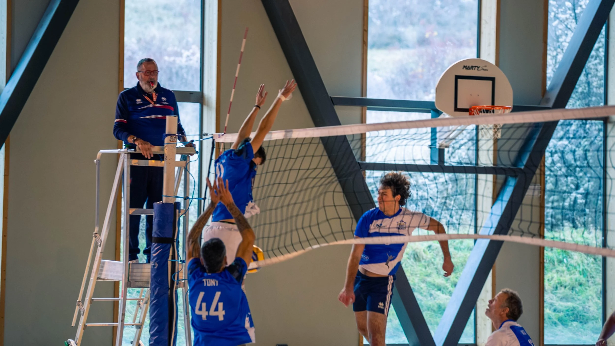 Joueur de volleyball en action, sautant pour attaquer le ballon lors d'un match en intérieur, avec un arbitre observant devant un fond de fenêtres avec vue sur la nature.
