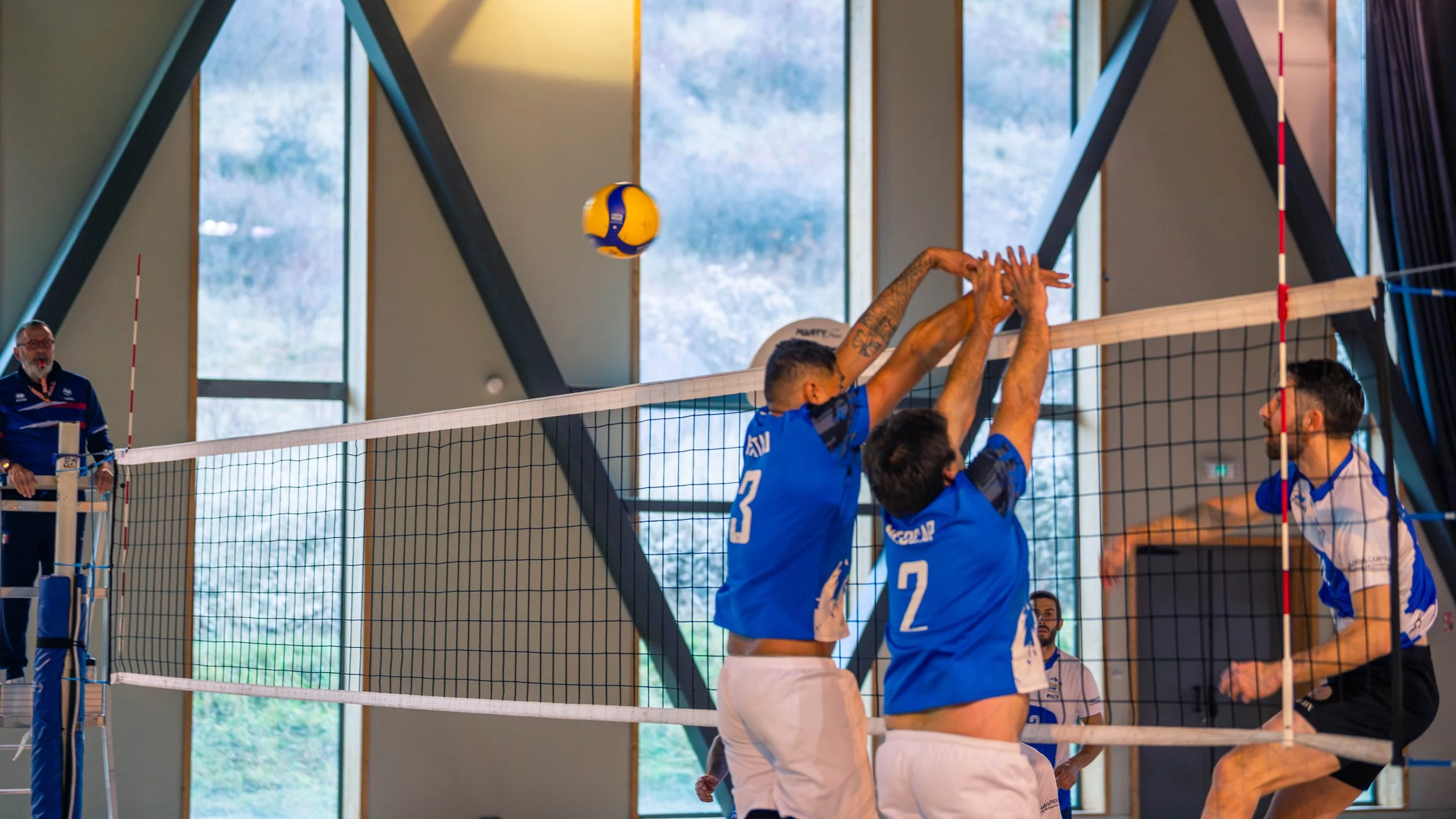 Une équipe de volleyball en action lors d'un match en salle, avec deux joueurs en bleu sautant pour bloquer le ballon au-dessus du filet, tandis qu'un autre joueur prépare une attaque.