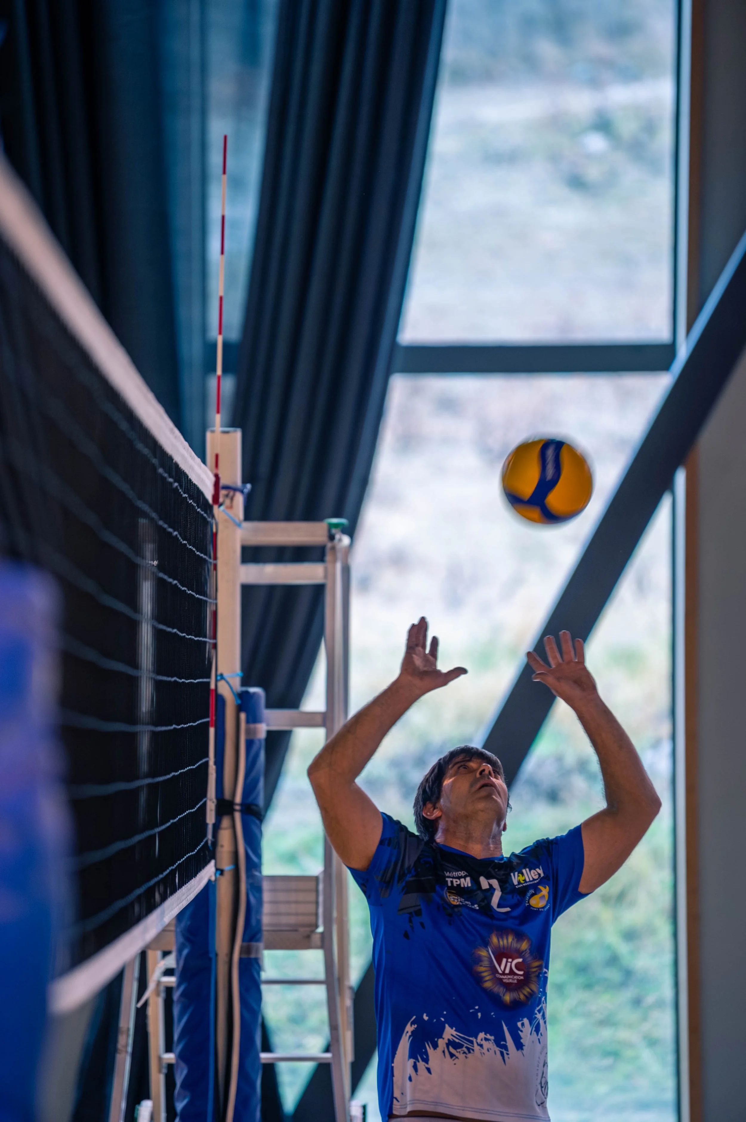 Un homme joue au volleyball en intérieur, prêt à recevoir ou retourner la balle. Il porte un maillot bleu avec des logos, et se trouve devant un mur avec un filet de volleyball. La scène est éclairée par de grandes fenêtres en arrière-plan.