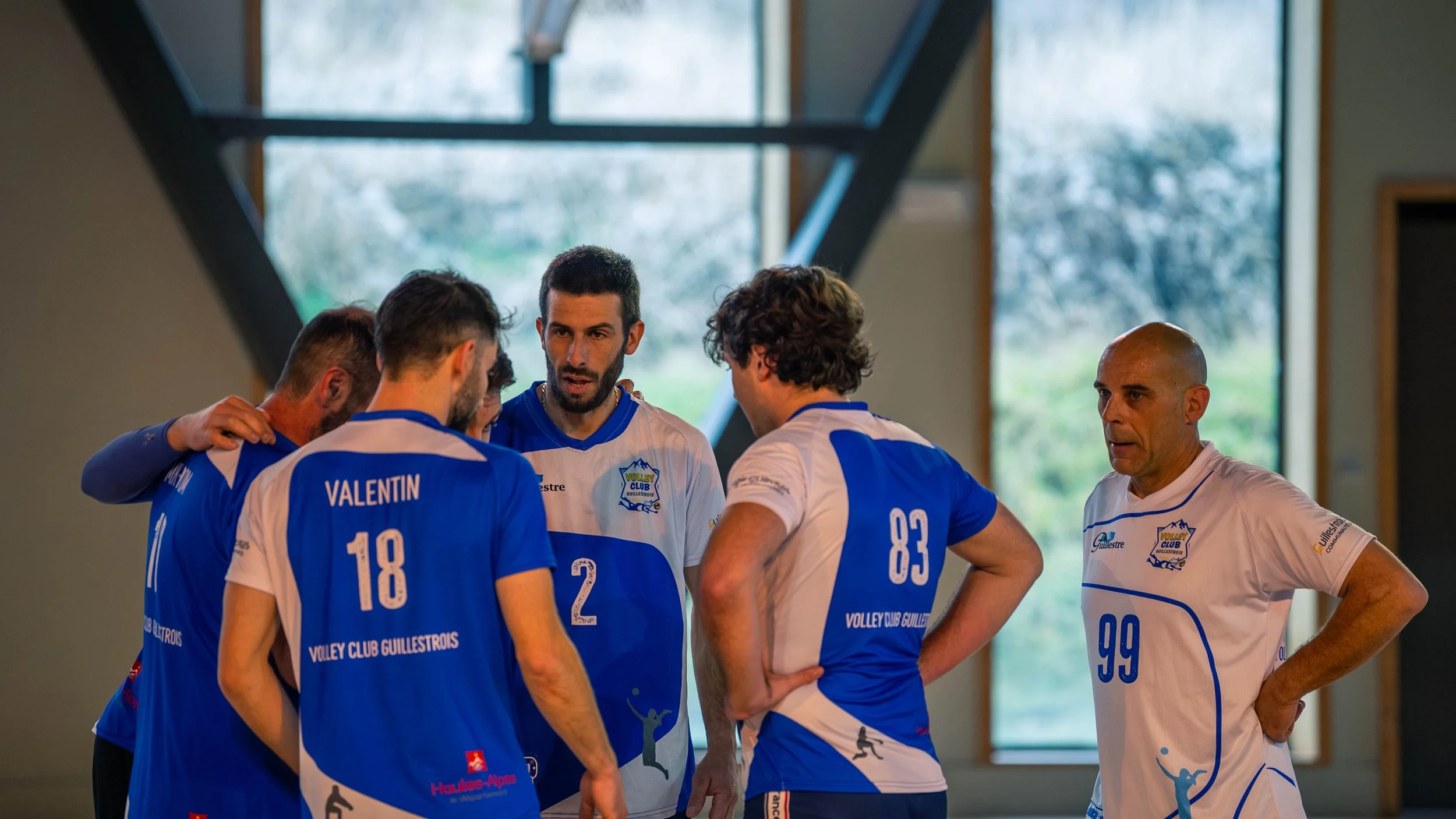L'équipe de volley-ball Guil lestroits en discussion lors d'un entraînement ou d'un match à l'intérieur, portant des maillots bleus et blancs avec le logo du club, dans une salle avec grandes fenêtres naturelles.