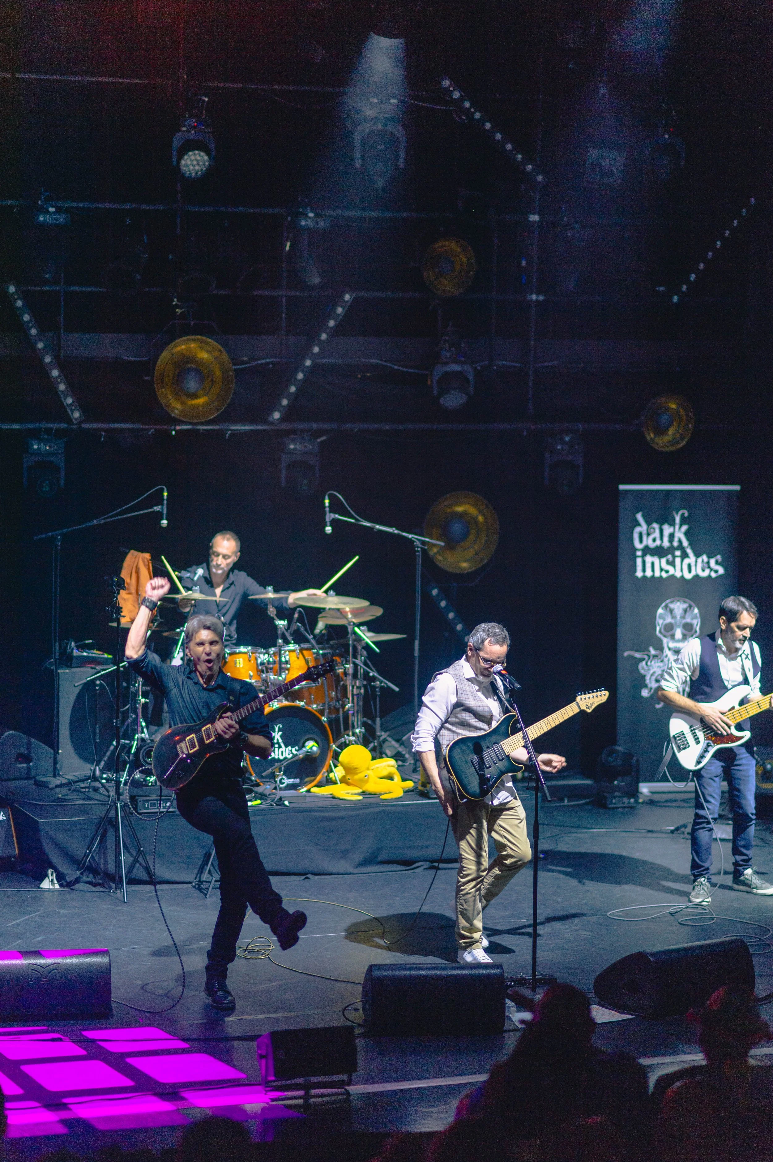 Un groupe de musiciens joue sur scène lors d'un concert, avec un batteur, deux guitaristes, et un bassiste, sous un éclairage de scène avec un décor noir et un panneau "dark insides" à l'arrière.