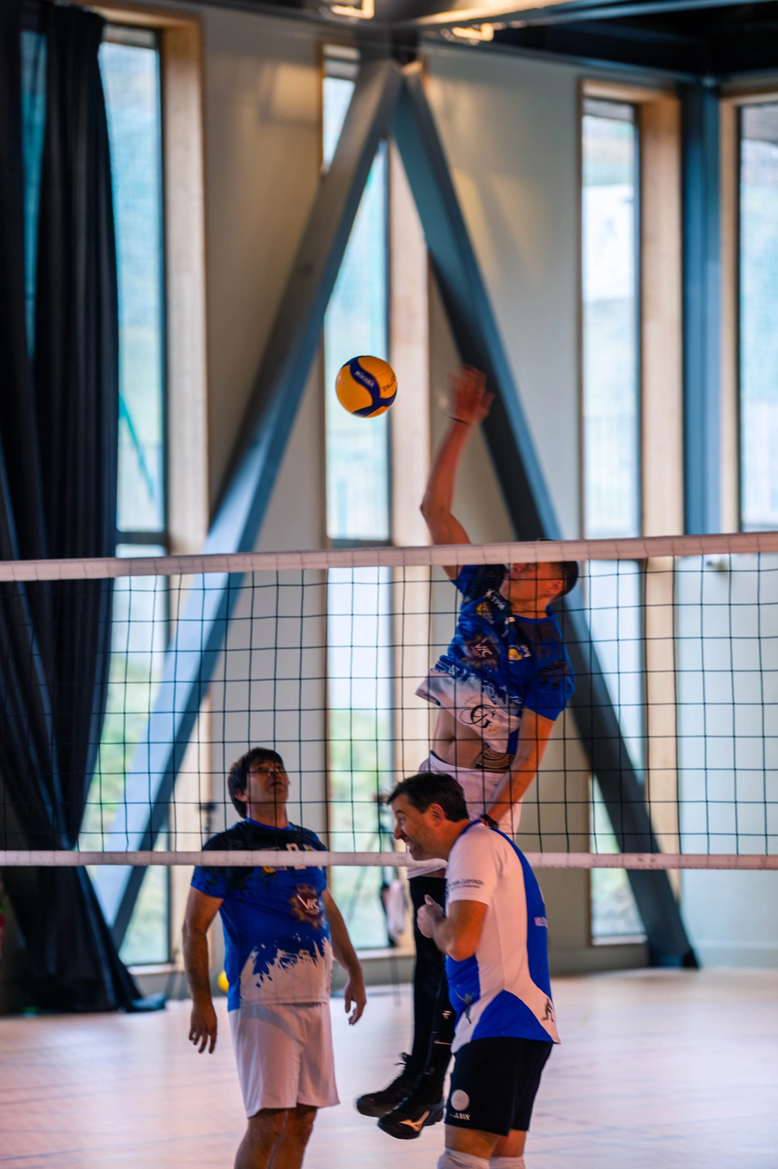 Un joueur de volley-ball en action sautant pour frapper le ballon au-dessus du filet, tandis que deux autres joueurs se tiennent en dessous, dans une salle de sport avec de grandes fenêtres et une structure en métal en arrière-plan.