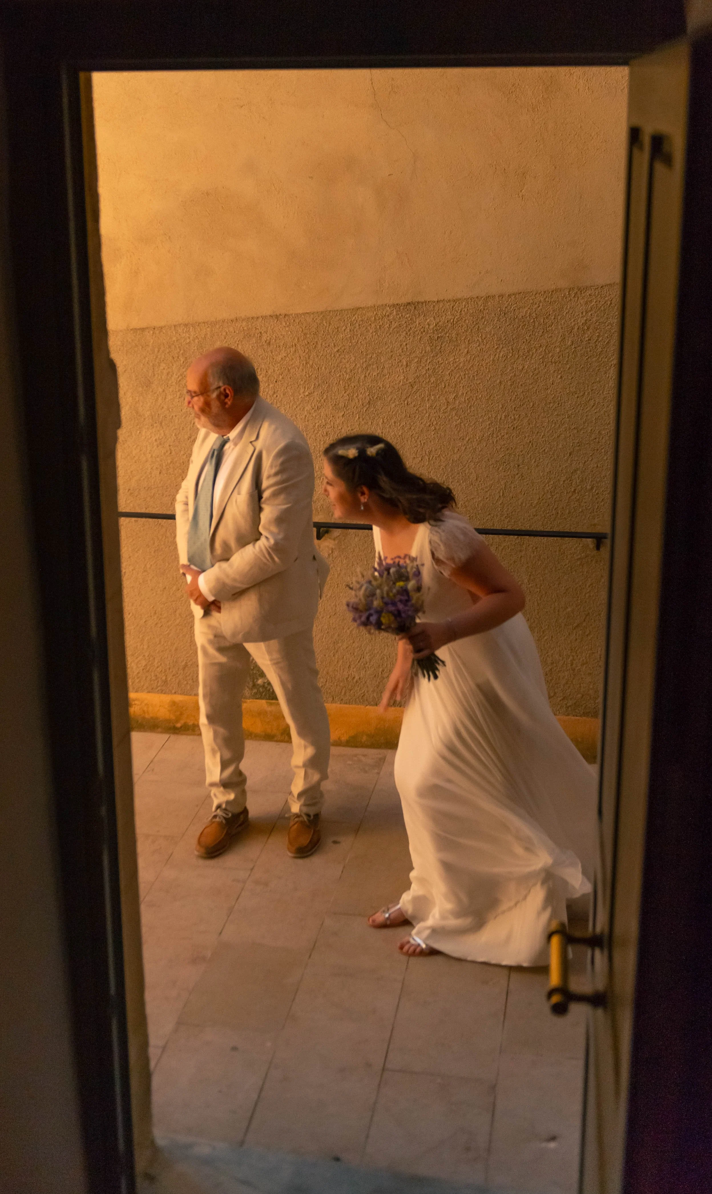 Une jeune femme en robe de mariée avec un bouquet de fleurs et un homme en costume blanc à l'extérieur, vus de derrière à travers une porte.