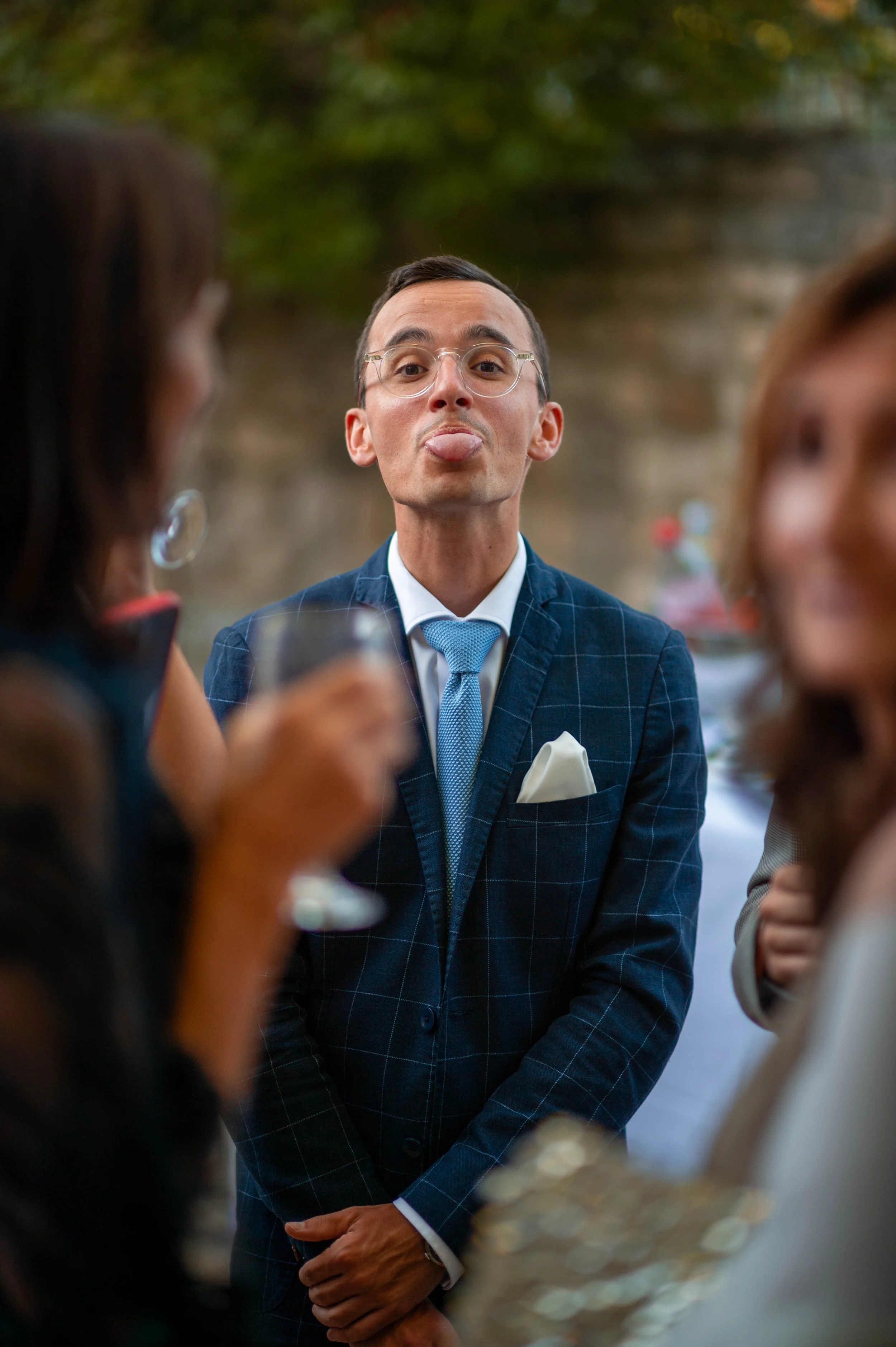 Homme en costume bleu avec une cravate, portant des lunettes, faisant une grimace avec la langue sortie lors d'une réunion sociale en extérieur.