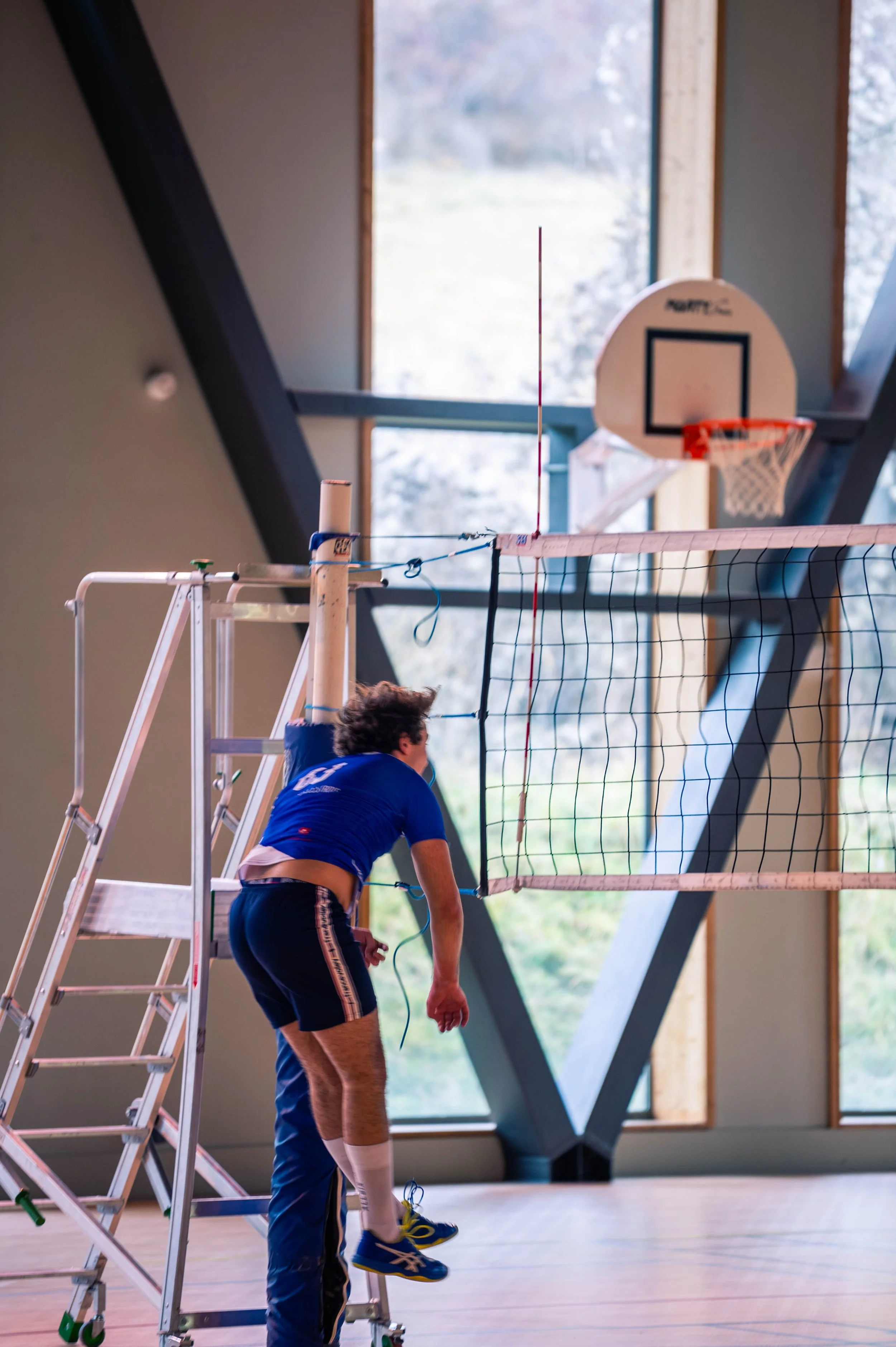 Un jeune homme en tenue sportive s'apprête à sauter lors d'un match de volleyball en intérieur, avec un panier de basketball en arrière-plan.