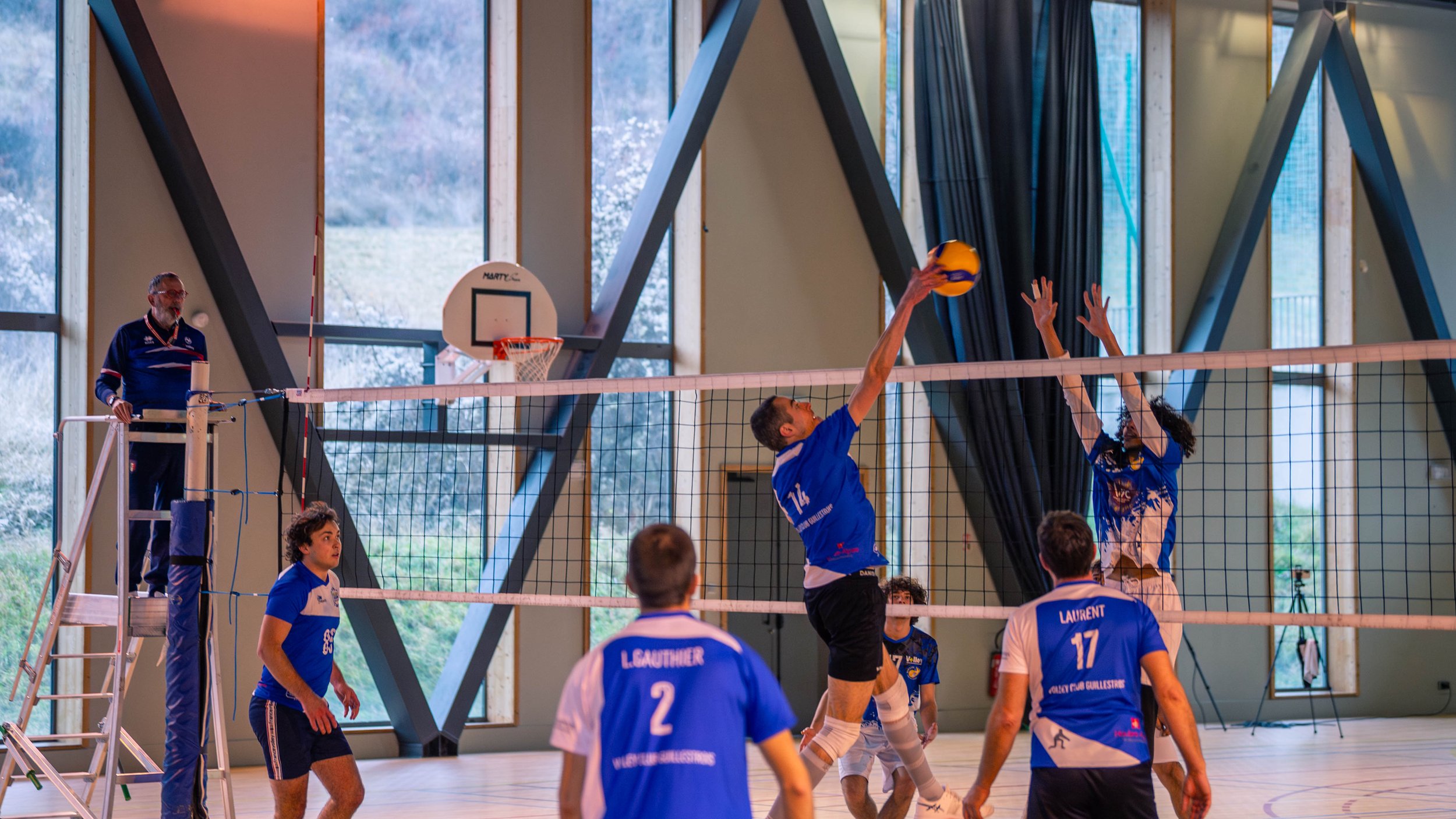 Une partie de volleyball en intérieur avec des joueurs en action, un arbitre sur une plateforme, et un filet au centre de l'image, avec des grands fenêtres en arrière-plan.