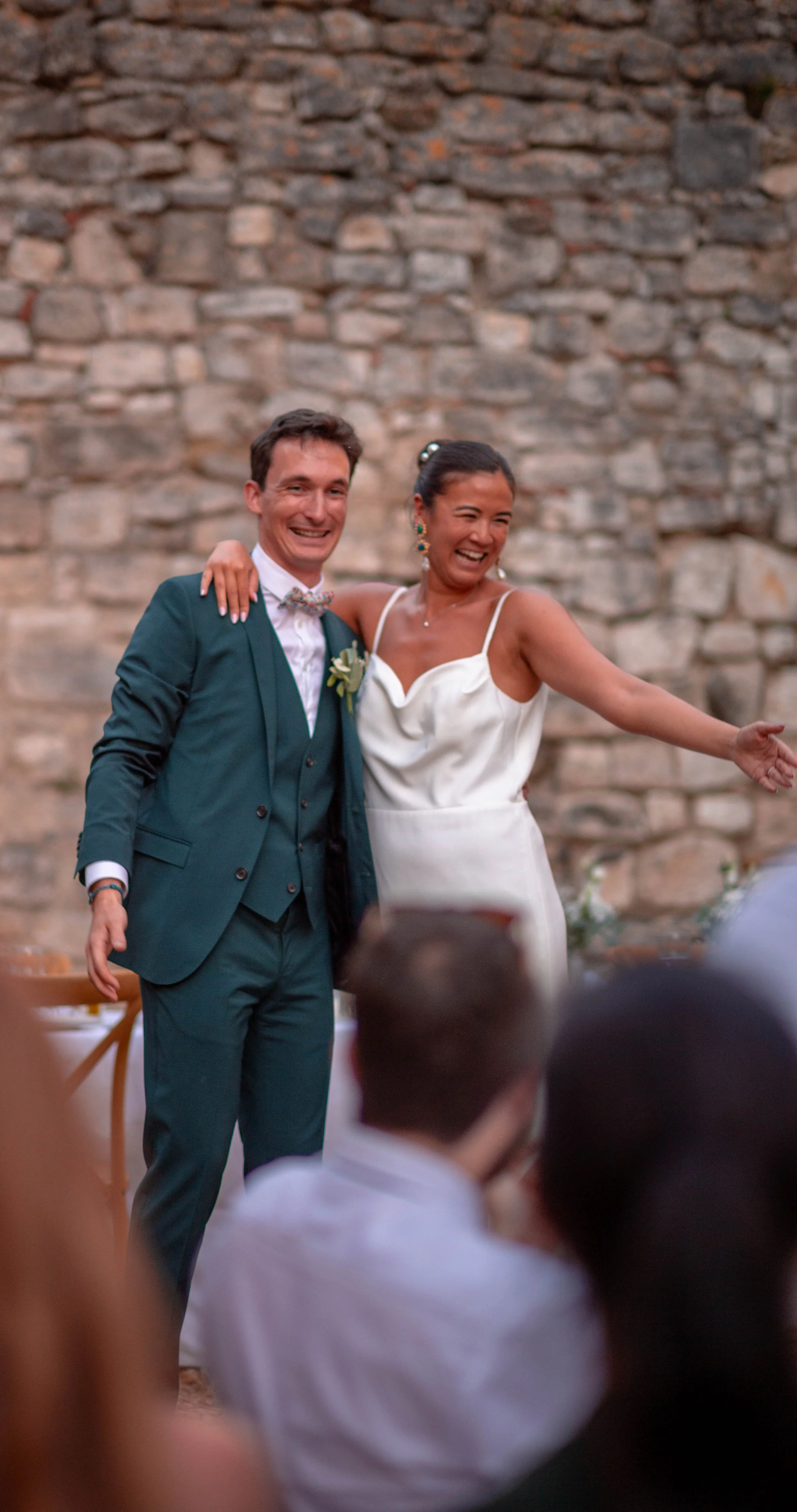 Deux personnes, un homme en costume vert et une femme en robe blanche, souriants lors d'un mariage, se tenant à côté d'une vieille paroi en pierre, en pleine célébration.
