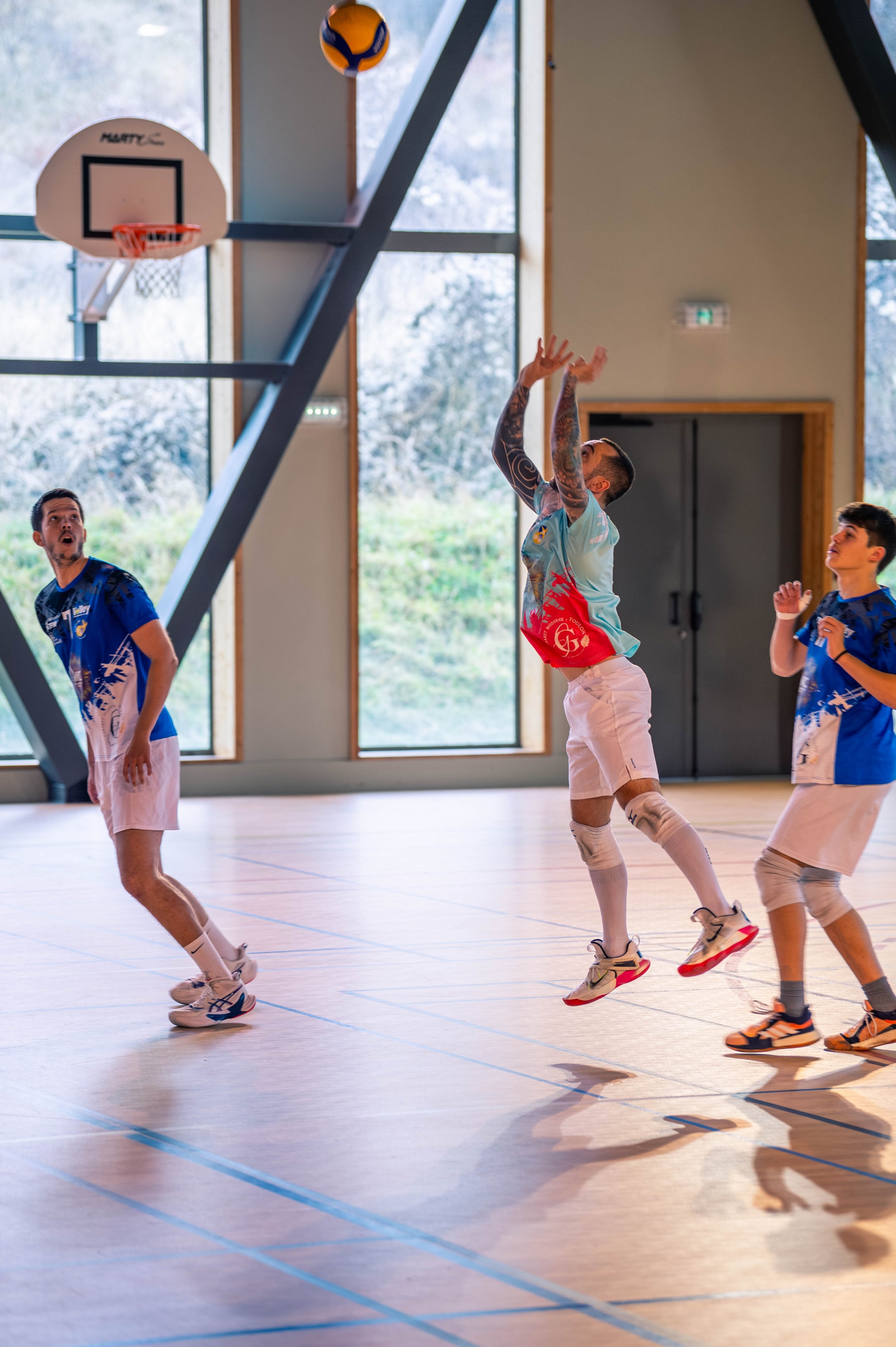 Des joueurs de volleyball en action à l'intérieur, avec un joueur sautant pour attraper la balle, dans un gymnase avec grandes fenêtres et un sol de sport en bois.