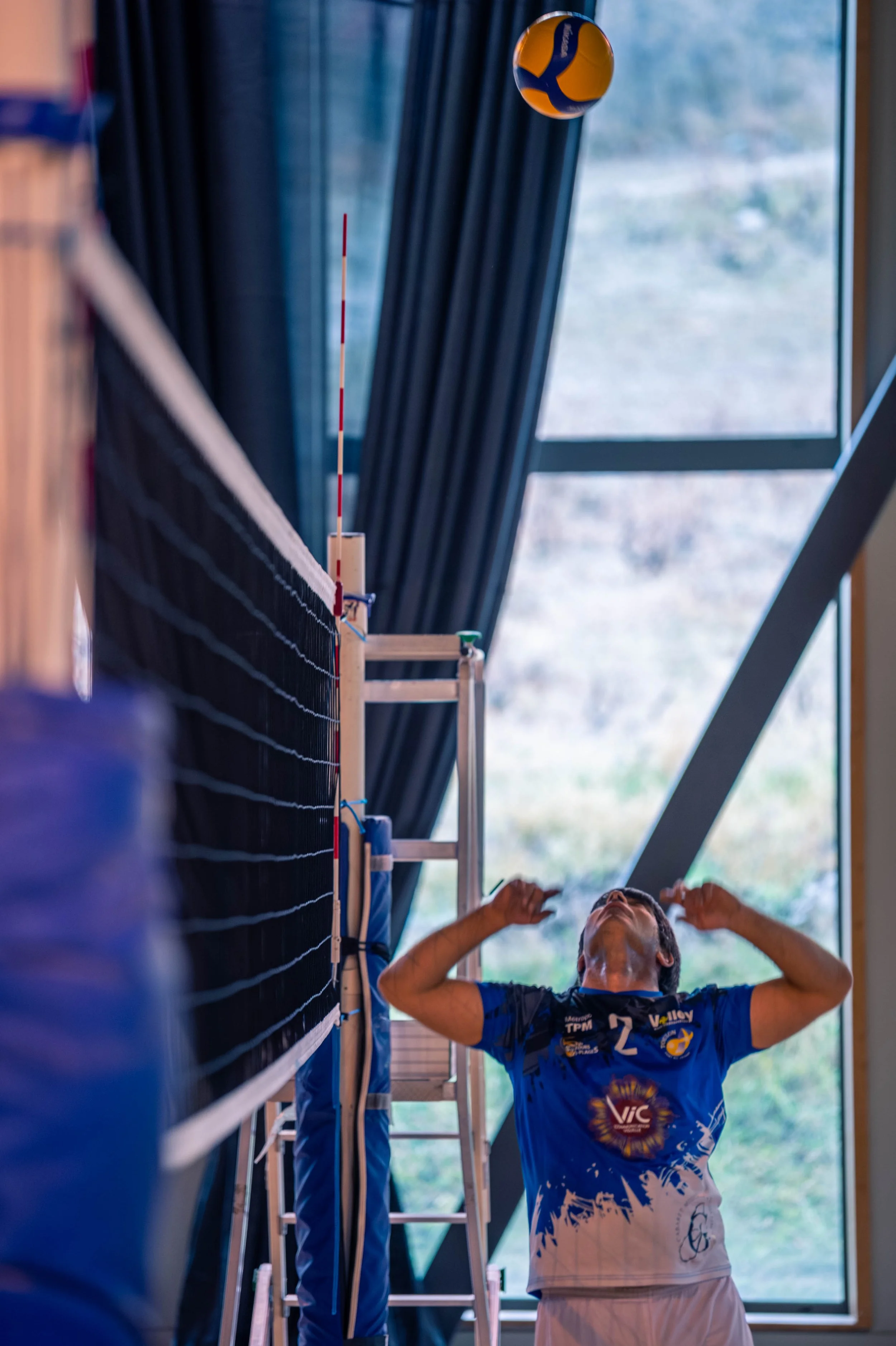 Un joueur de volleyball en train de se préparer à réceptionner la balle dans une salle intérieure, avec un filet de volleyball et une fenêtre en arrière-plan.