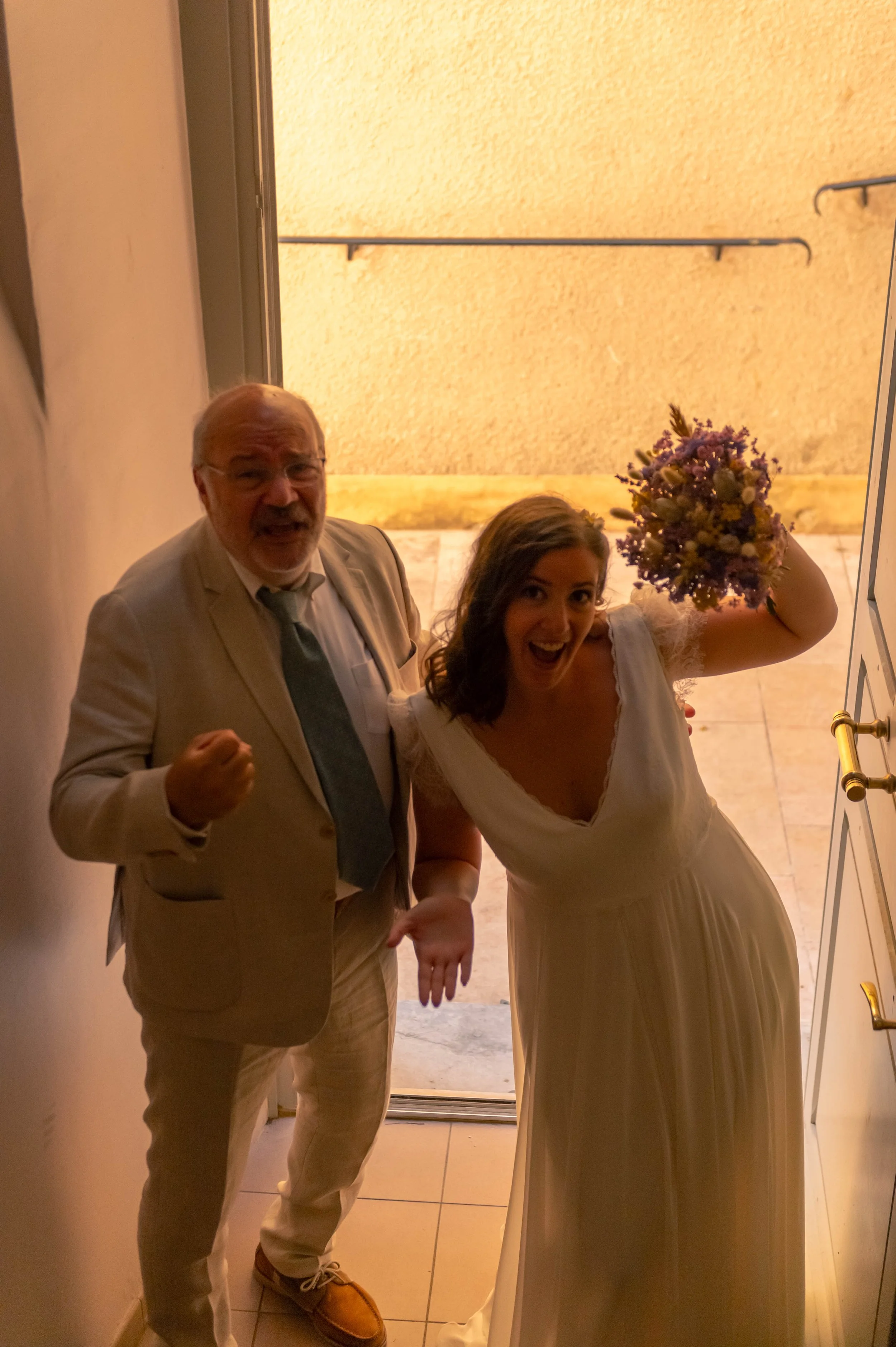 Une femme en robe de mariage heureuse tenant un bouquet de fleurs, et un homme en costume criant de joie, posant dans l'entrée d'une pièce.