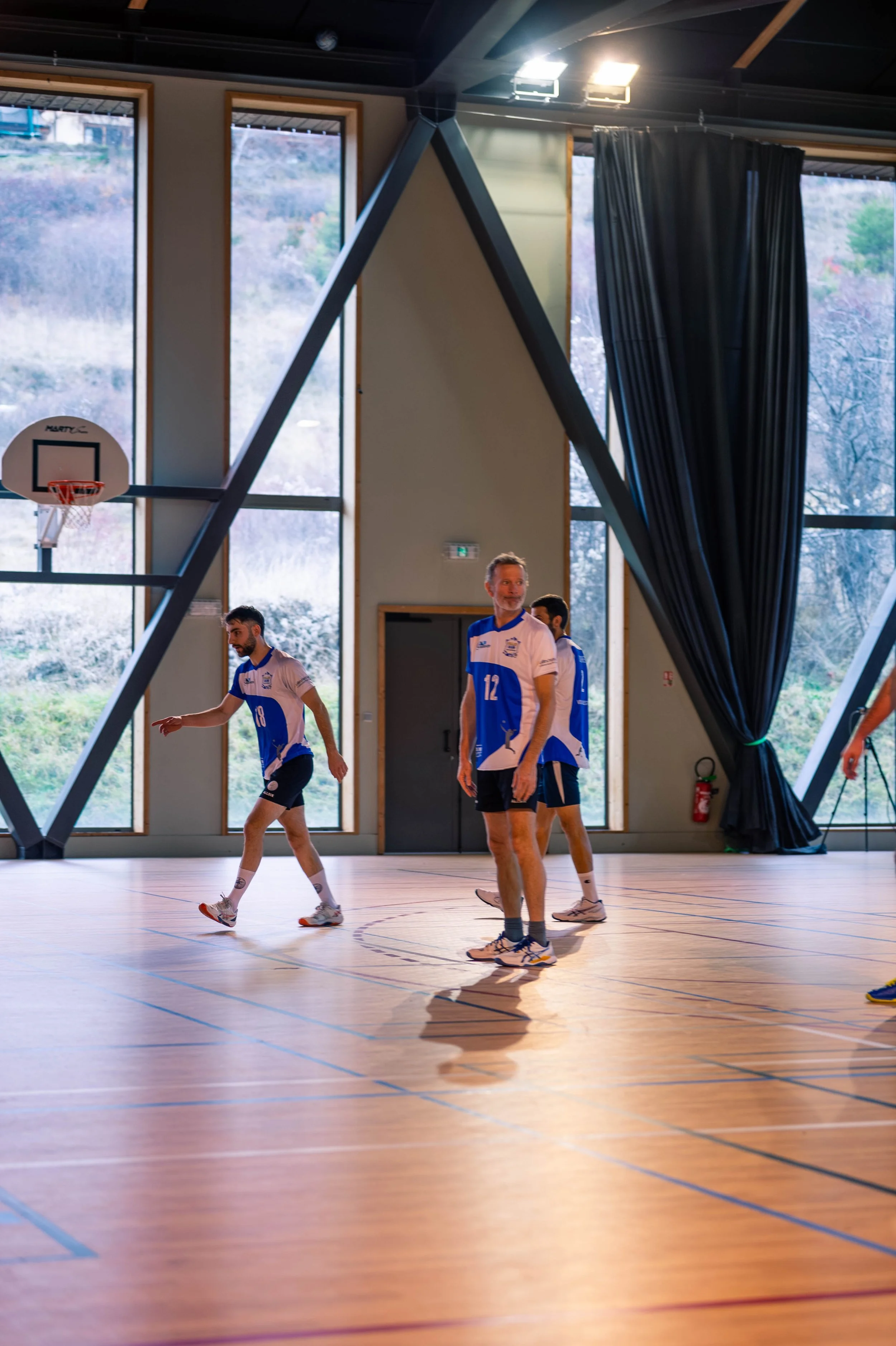 Joueurs de volleyball dans un gymnase avec murs en verre et terrain en bois.