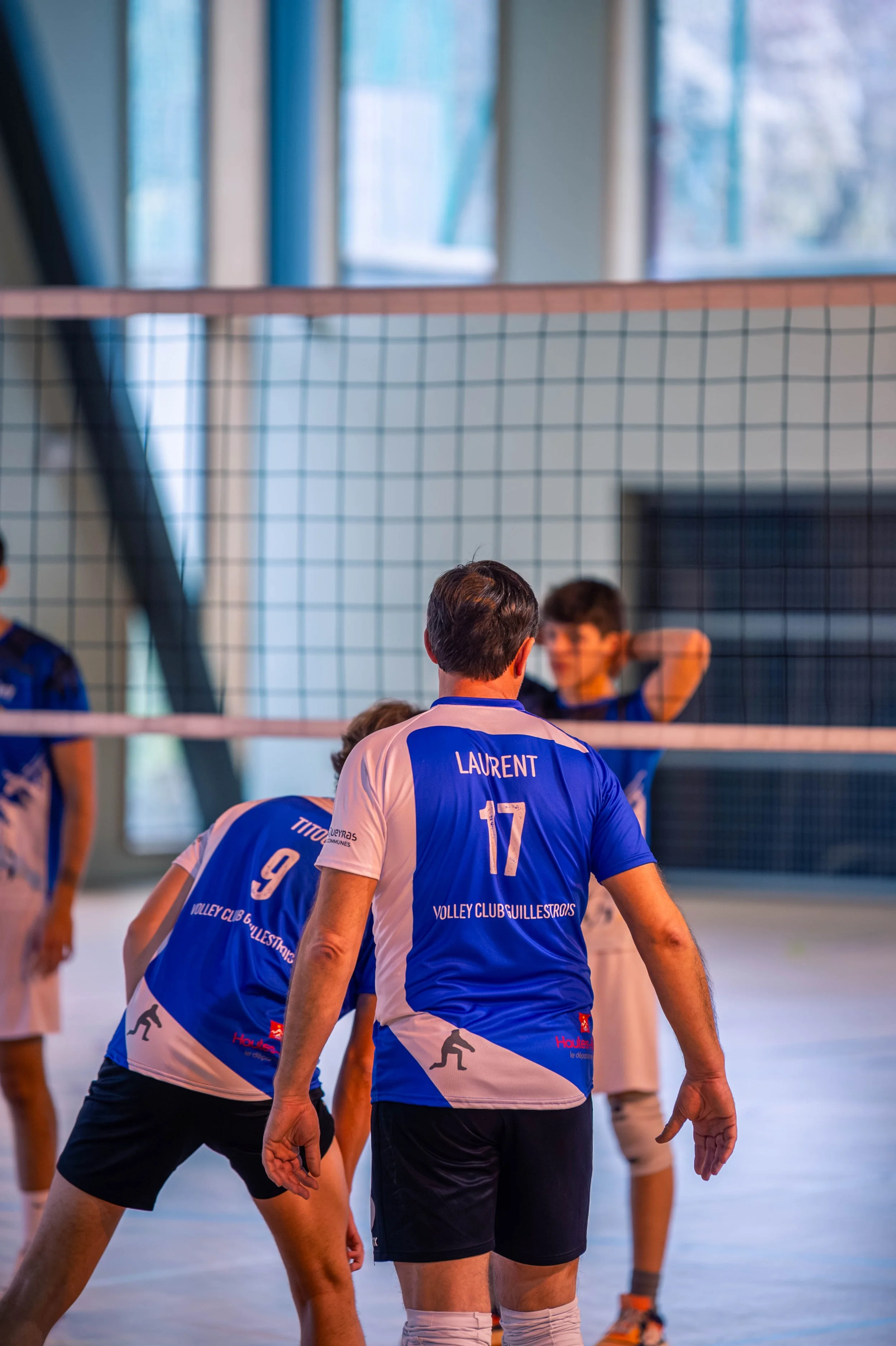 Joueurs de volleyball en tenue bleue, en train de jouer dans une salle d'escalade, vue de dos, avec un filet au centre et des fenêtres en arrière-plan.