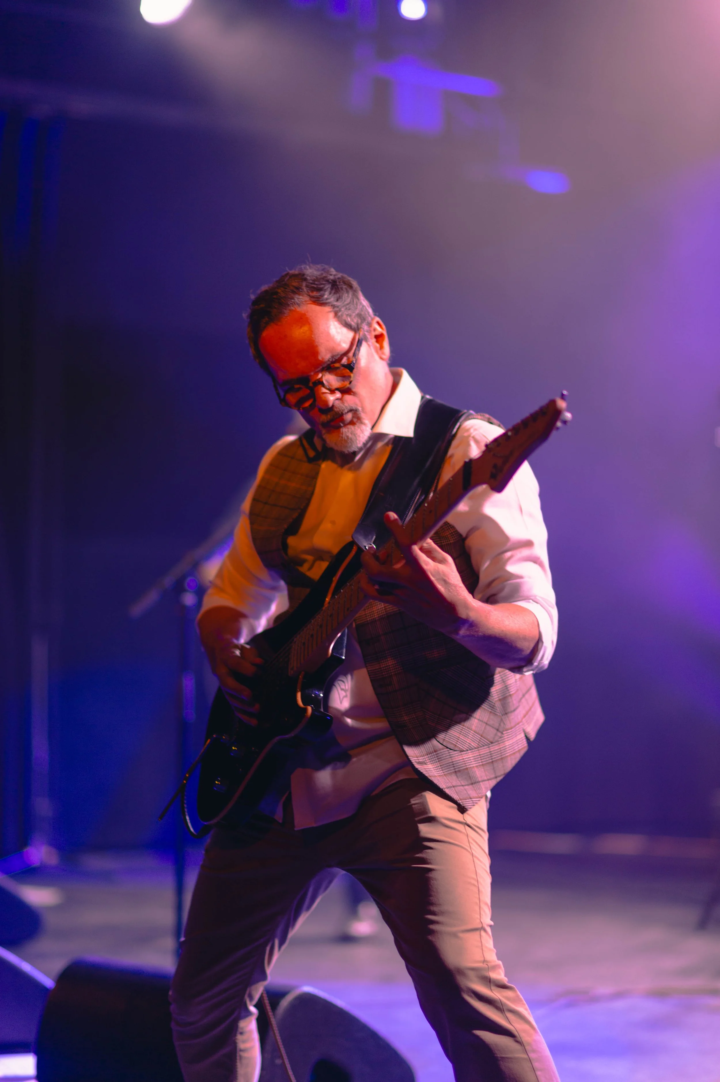 Un homme joue de la guitare électrique sur scène, éclairé par des lumières violettes et bleues, lors d'une performance musicale.