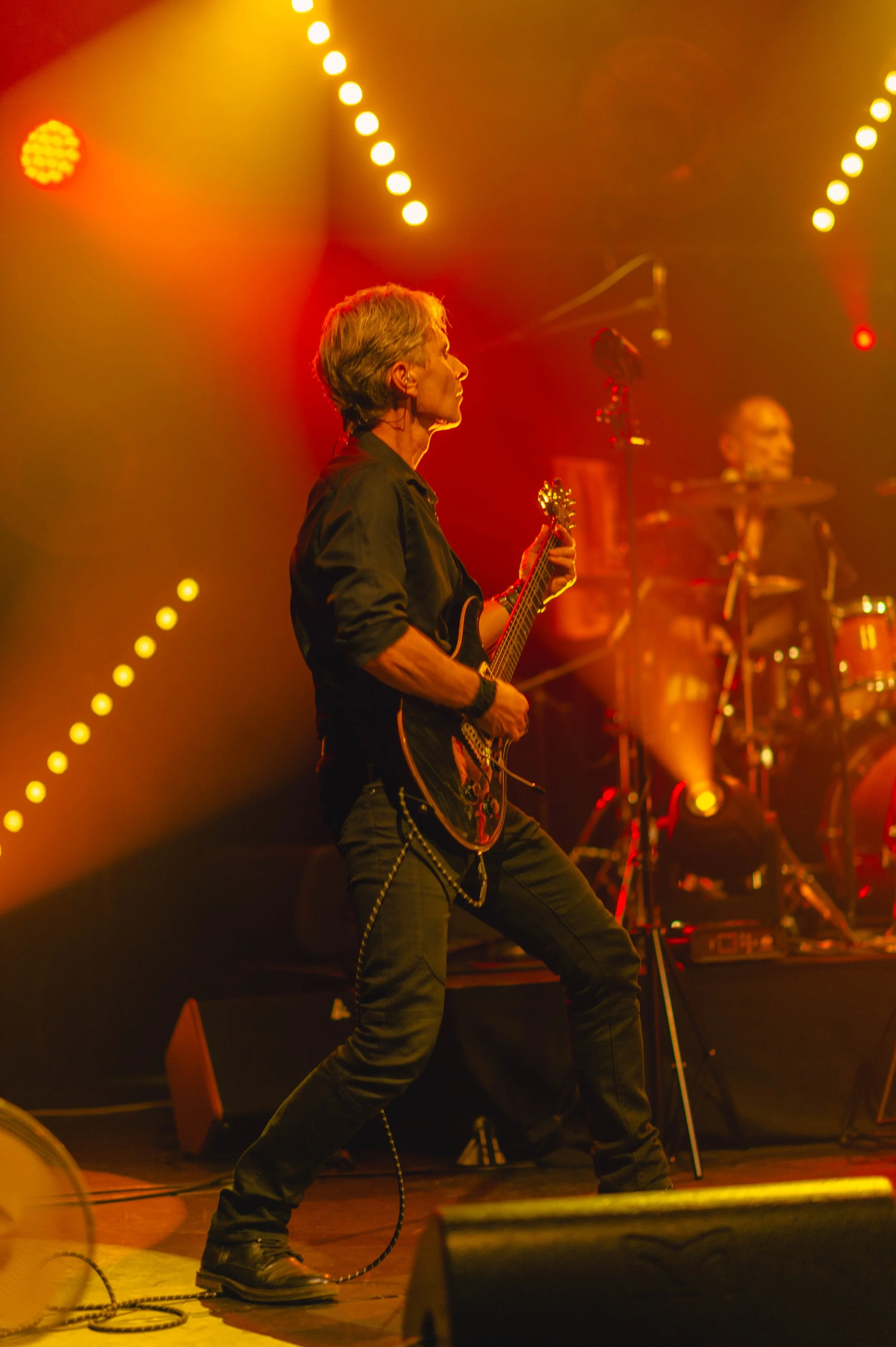 Un musicien jouant de la guitare électrique sur scène lors d'un concert, avec un batteur en arrière-plan, sous des lumières chaudes et colorées.