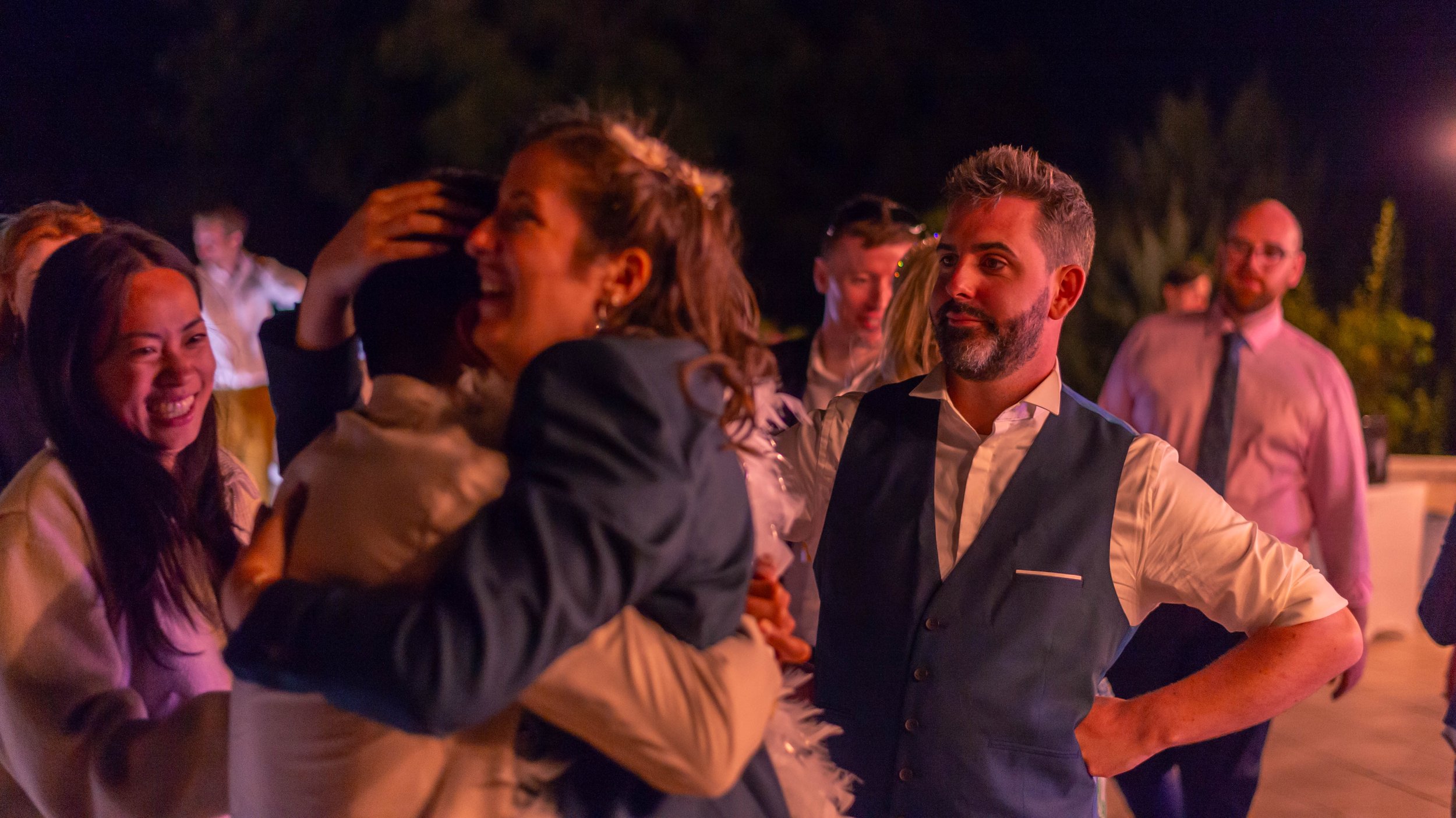 Groupe de personnes souriantes et riantes lors d'une soirée ou fête en plein air, éclairée par des lumières colorées, avec un homme parée d'un veston sur une chemise blanche, entouré d'autres personnes en arrière-plan.