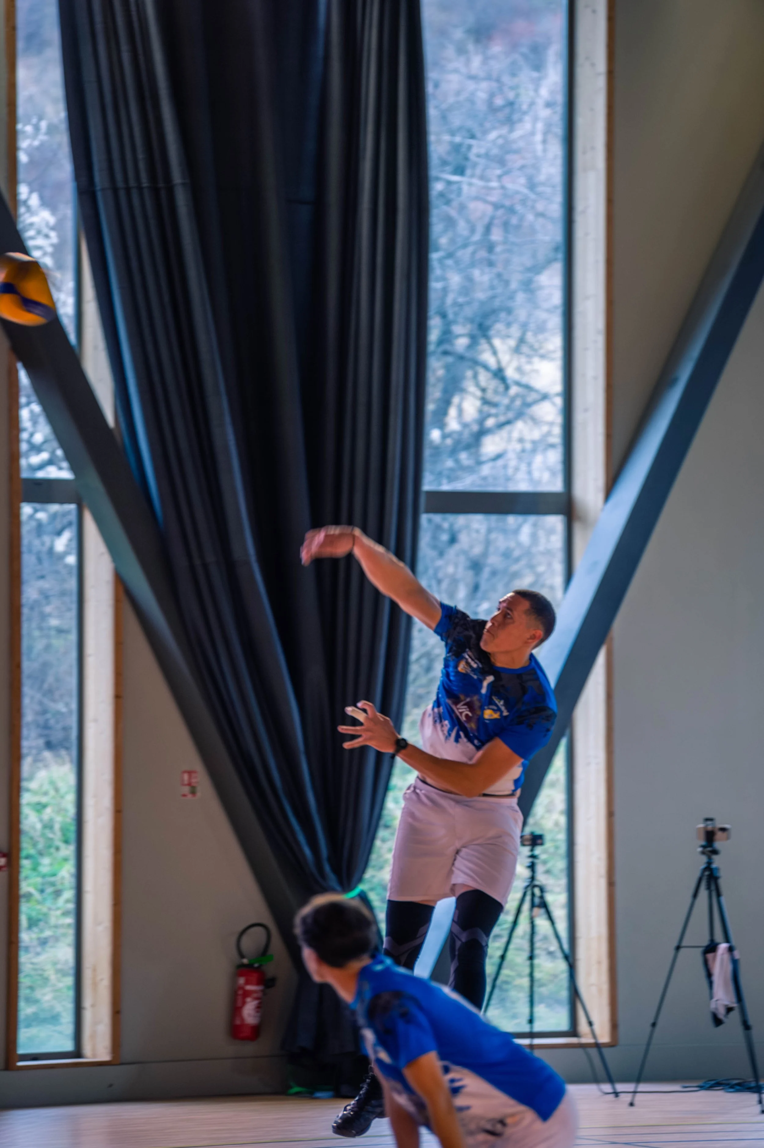 Un homme en tenue sportive joue au volleyball en intérieur, avec un jeune garçon au premier plan. Des appareils photo sont posés sur des trépieds dans la pièce, et de grandes fenêtres laissent entrer la lumière naturelle.