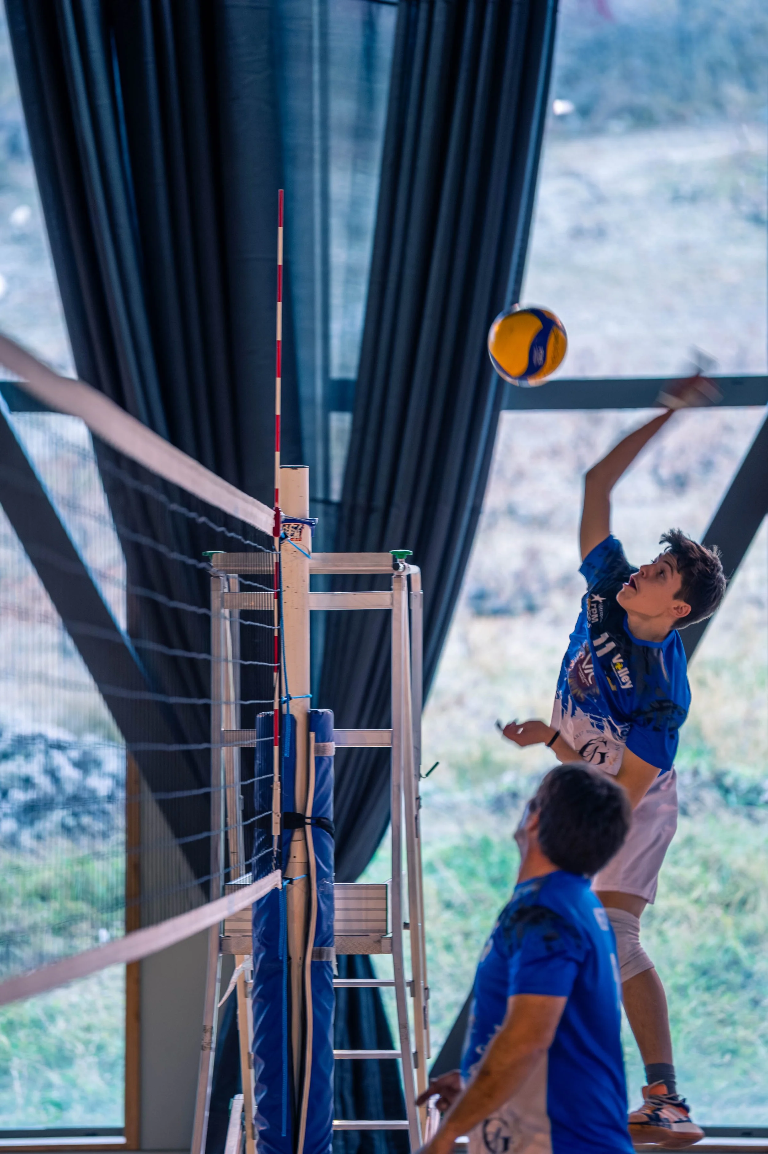 Un jeune joueur de volley-ball en pleine action lors d'un match intérieur, sa main levée pour frapper le ballon en vol, deux autres personnes en arrière-plan dans un environnement sportif.