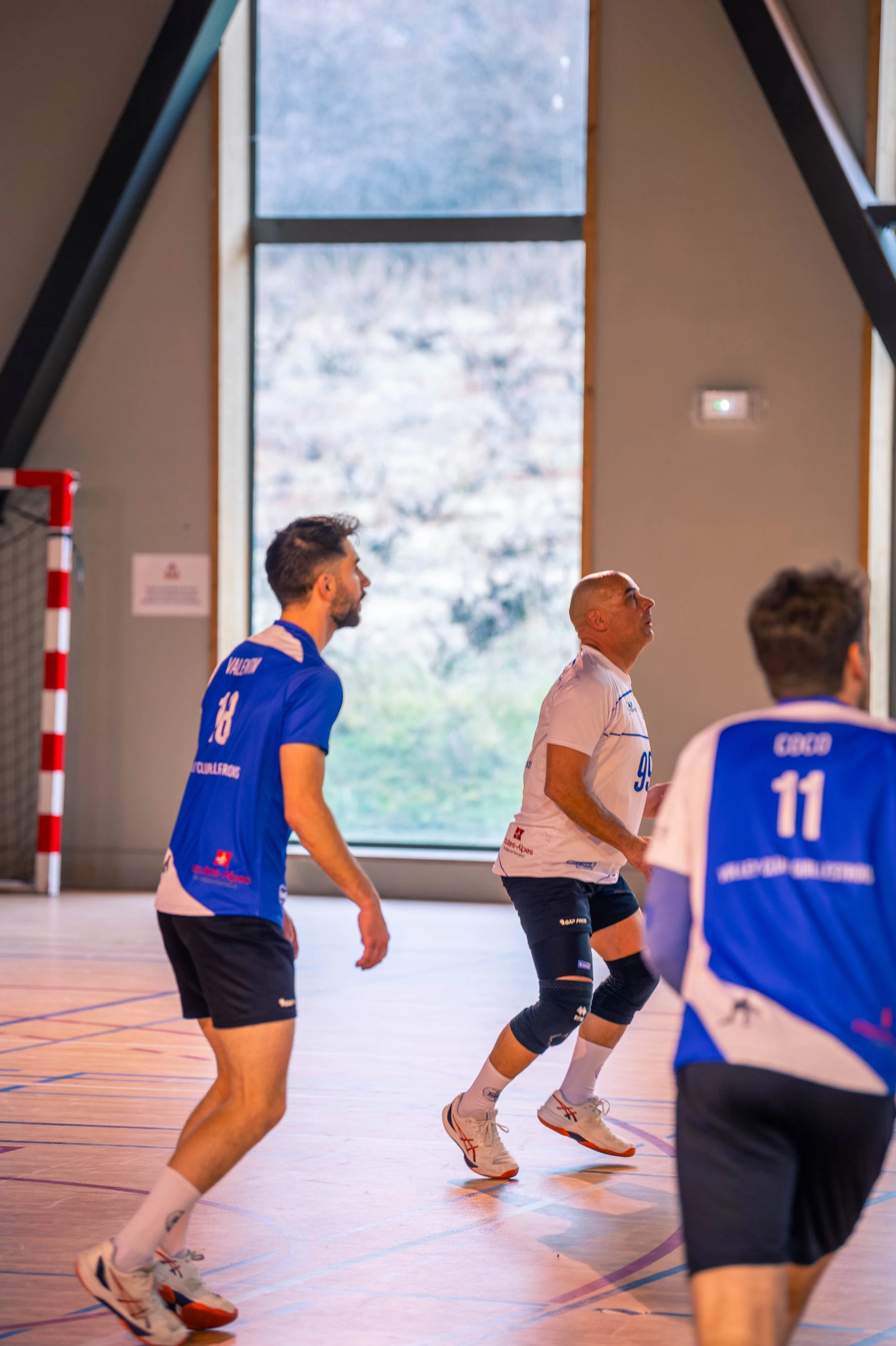 Joueurs de volleyball en intérieur s'entraînant ou jouant, portant des maillots bleus et blancs, dans une salle avec un grand fenêtre en arrière-plan laissant passer la lumière naturelle.