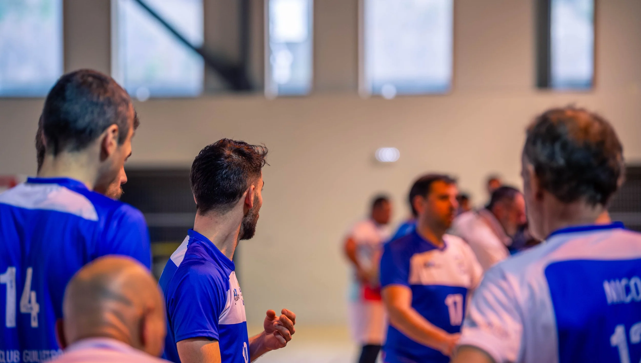 Une équipe de handball en tenue bleue, en réunion dans un gymnase, avec des fenêtres en haut et un mur beige en arrière-plan.