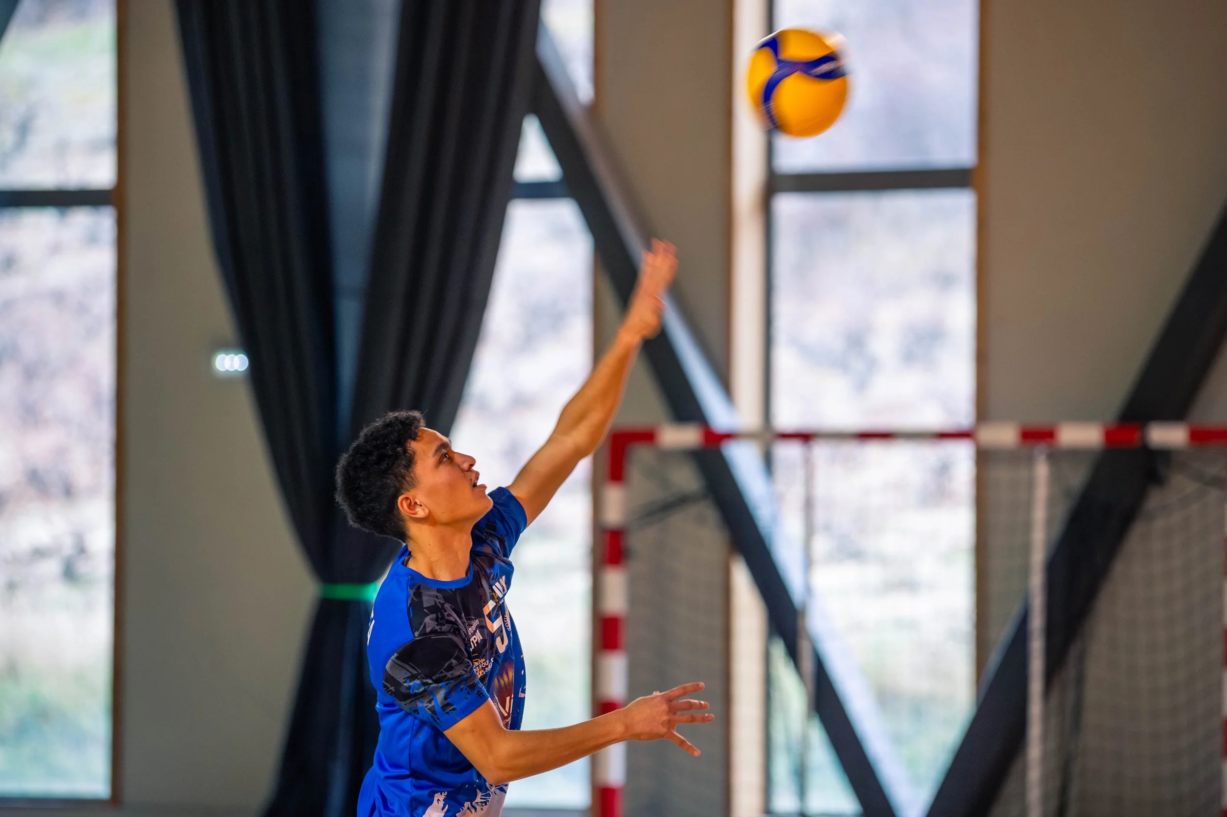 Une femme joue au volley-ball en intérieur, sautant pour frapper le ballon avec son bras tendu, dans une salle avec de grandes fenêtres et des rideaux noirs.