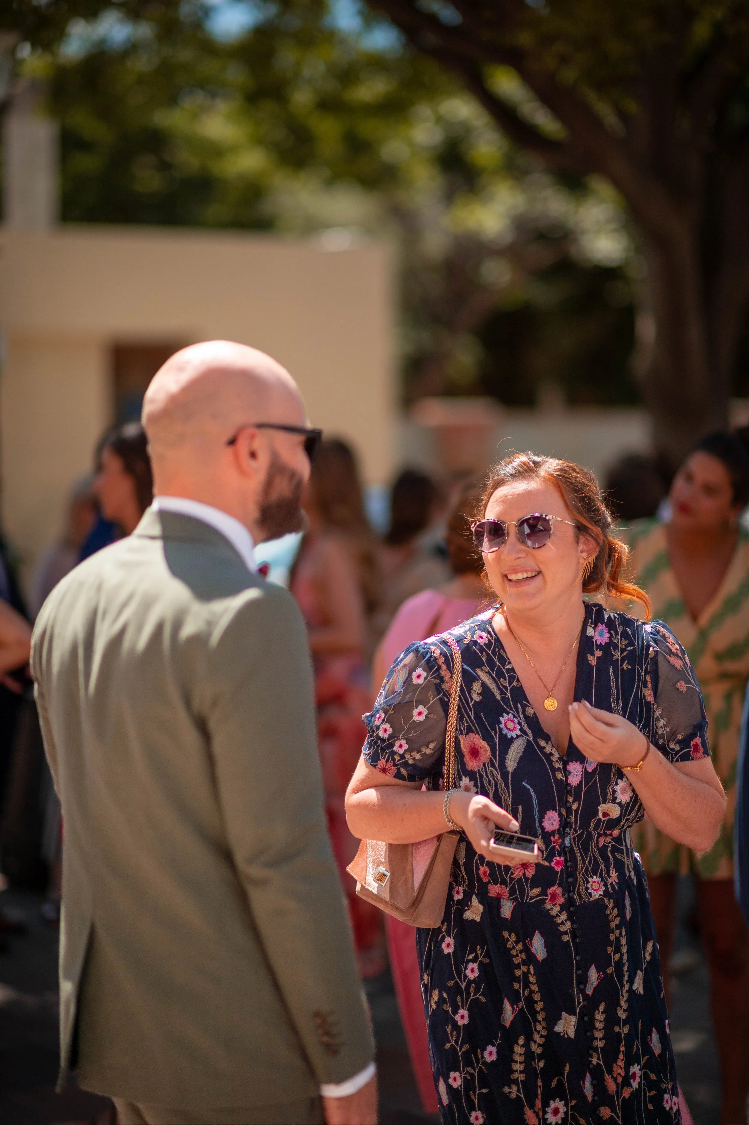 Deux personnes souriantes en conversation lors d'un événement extérieur en journée, entourées d'autres invités, avec des arbres en arrière-plan.