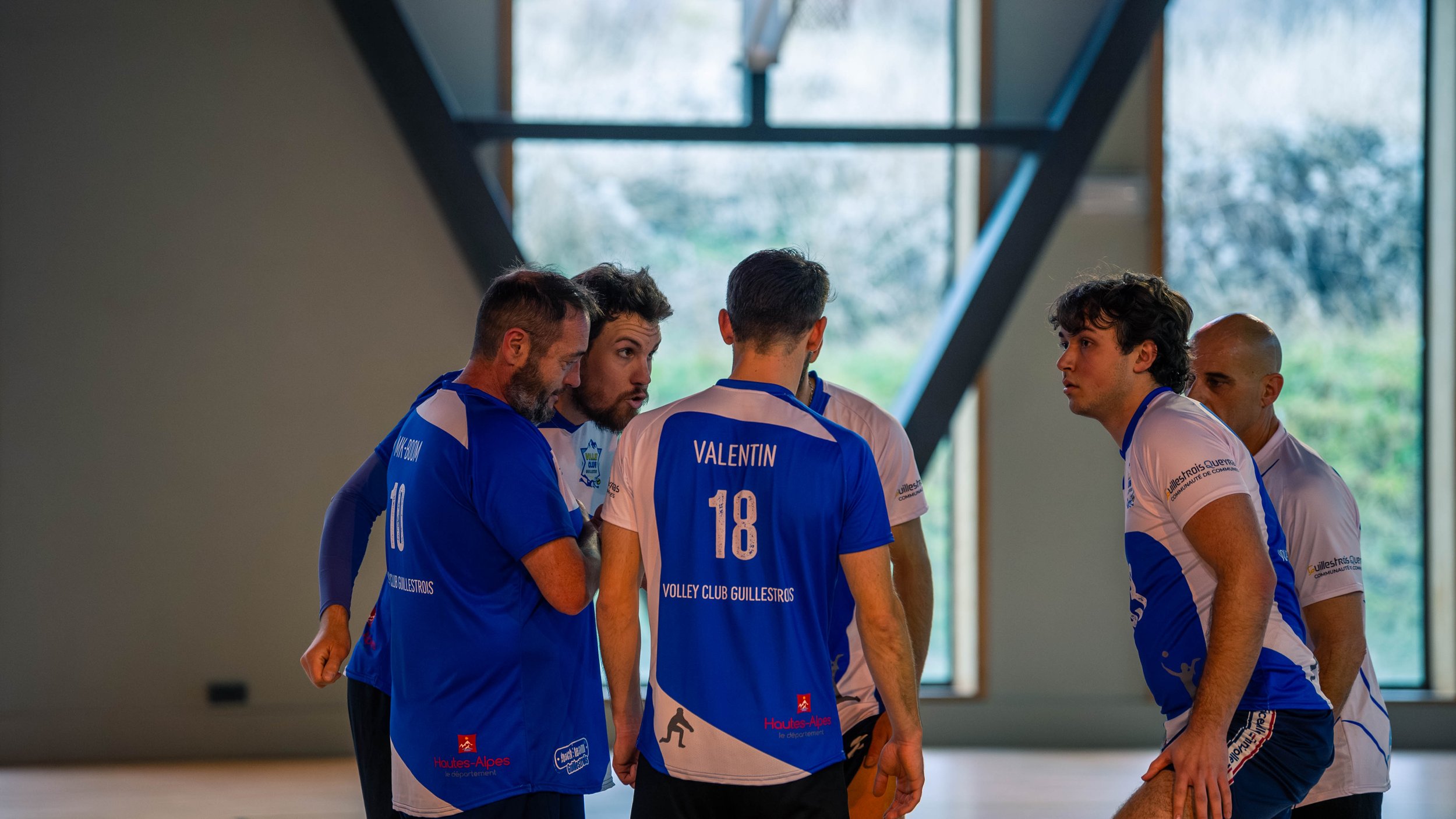 Une équipe de volleyball en discussion lors d'un entraînement ou d'un match dans une salle intérieure. Les joueurs portent des maillots bleus et blancs avec le nom 'Valentin' et le numéro '18' visible.