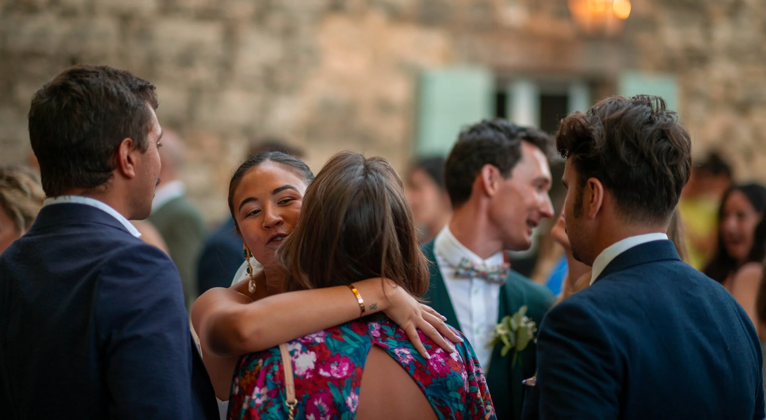 Groupe de personnes en conversation lors d'une fête ou d'un événement social, dans un espace intérieur avec mur en pierre. Des hommes en costumes et des femmes se décrivent et échangent chaleureusement.