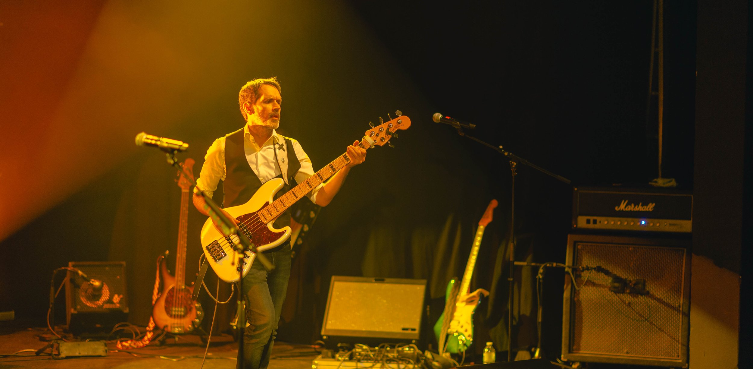 Musicien jouant de la basse électrique sur scène, entouré de matériel audio et de guitares, sous un éclairage jaune-orange, avec un amplificateur Marshall visible à droite.