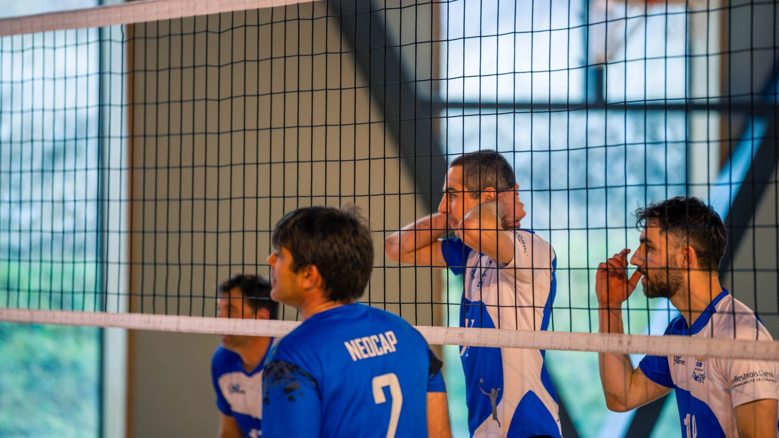 Joueurs de volleyball dans un gymnase, un d'eux se prépare à servir, portant un maillot bleu avec le numéro 2, en conversation et en attente pendant un match.
