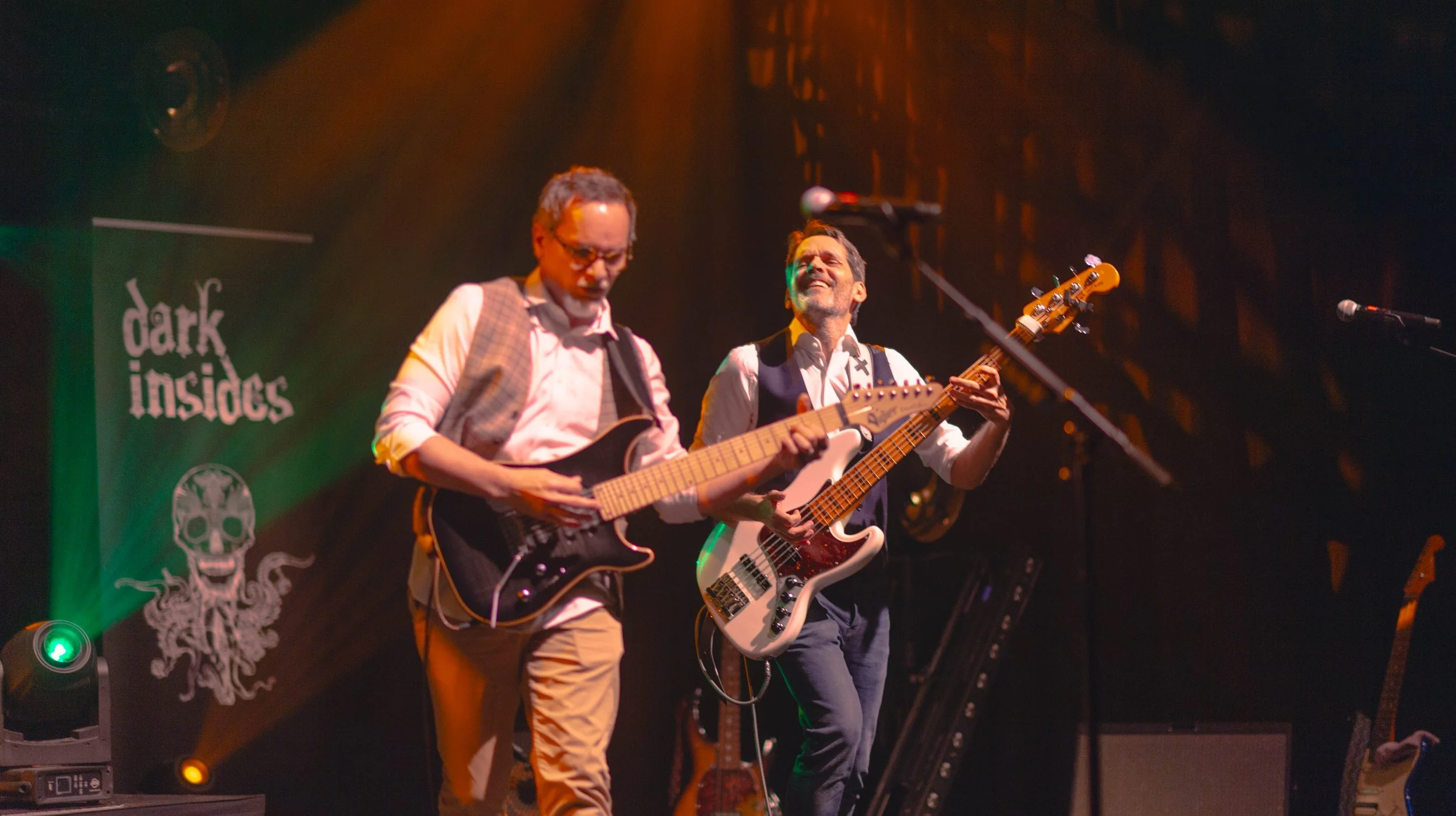 Deux musiciens jouent de la guitare lors d'un concert, avec un fond sombre et un panneau indique 'dark insides'.