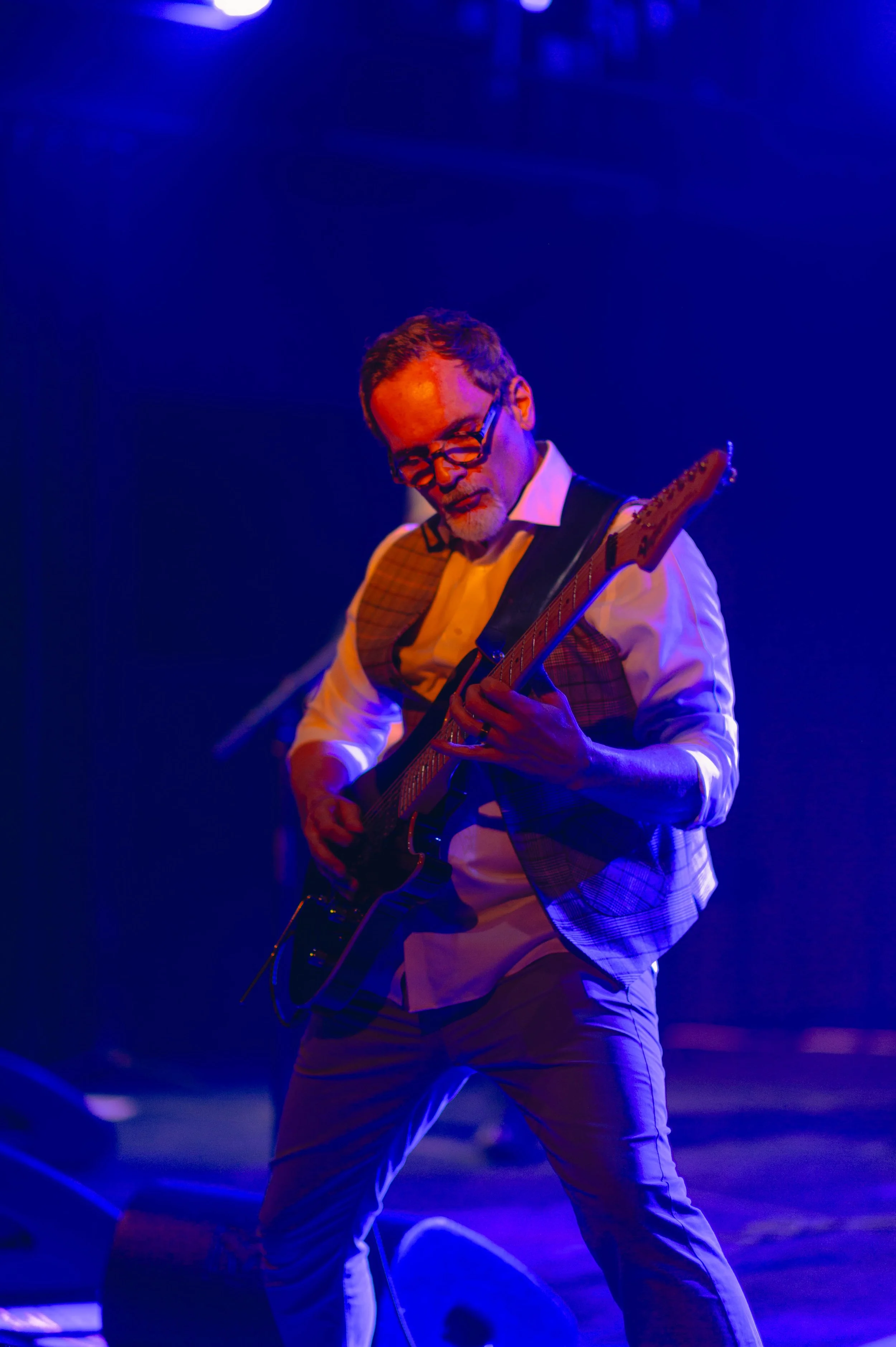 Un homme jouant de la guitare électrique lors d'une performance sur scène sous un éclairage bleu, portant une chemise blanche, un veston à carreaux, et des lunettes.