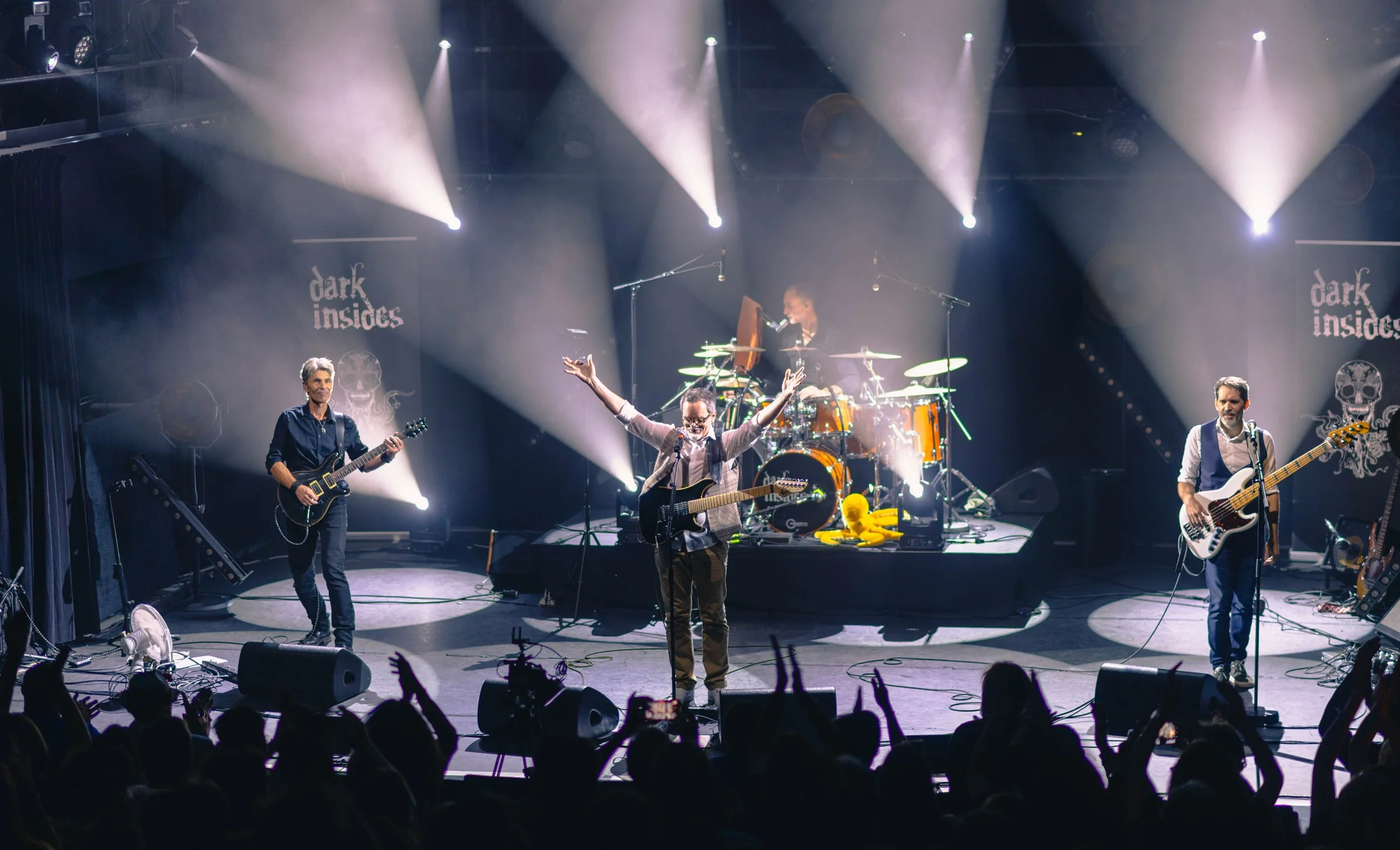 Un groupe de musiciens en concert avec le logo "dark insides" en arrière-plan, scène éclairée par des projecteurs, public en admiration.