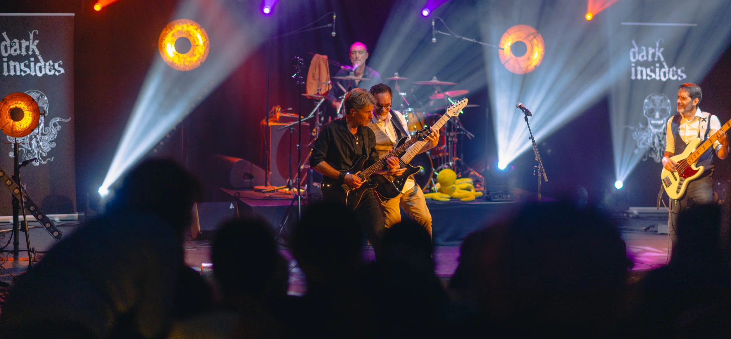 Groupe de musiciens jouant sur scène lors d'un concert, avec éclairages colorés et écrans en arrière-plan affichant 'dark insides' et des symboles.