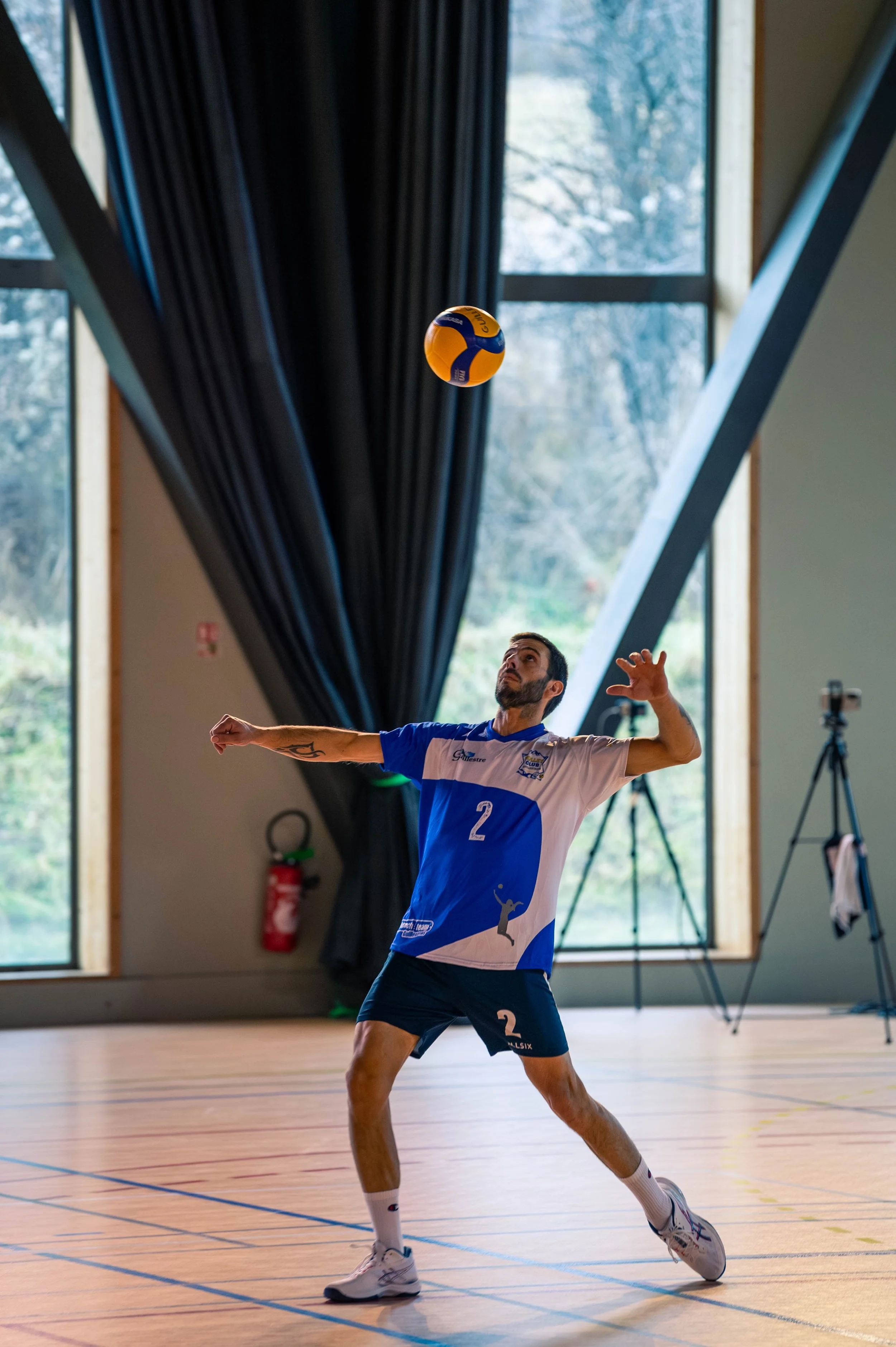 Un joueur de volleyball en intérieur, portant un maillot bleu et blanc avec le numéro 2, se prépare à recevoir ou à frapper une balle de volley orange et bleue.