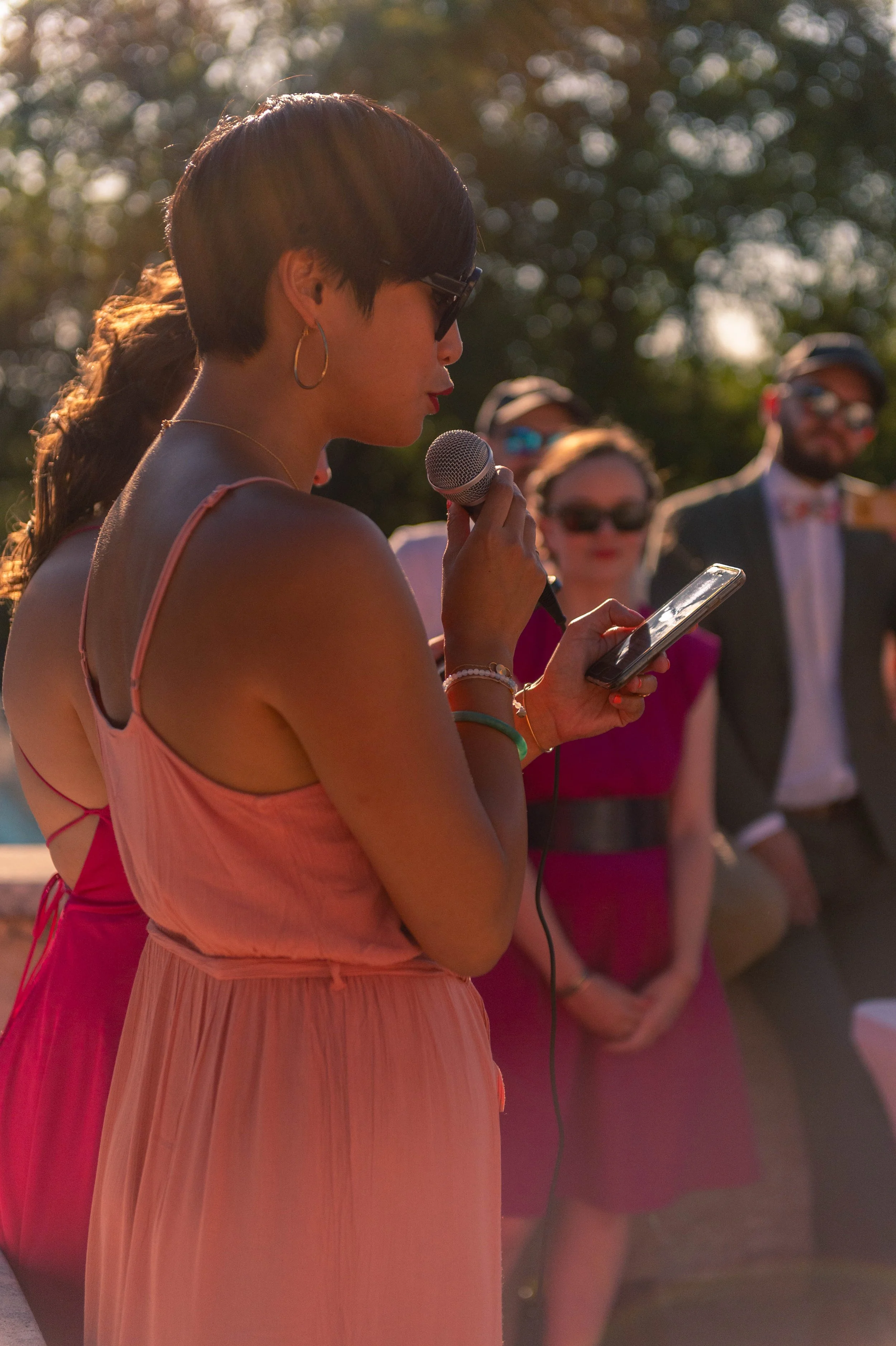 Une femme, portant des lunettes de soleil et une robe rose, tient un microphone et lit quelque chose sur son téléphone lors d'un événement en plein air en soirée, avec d'autres personnes en arrière-plan.