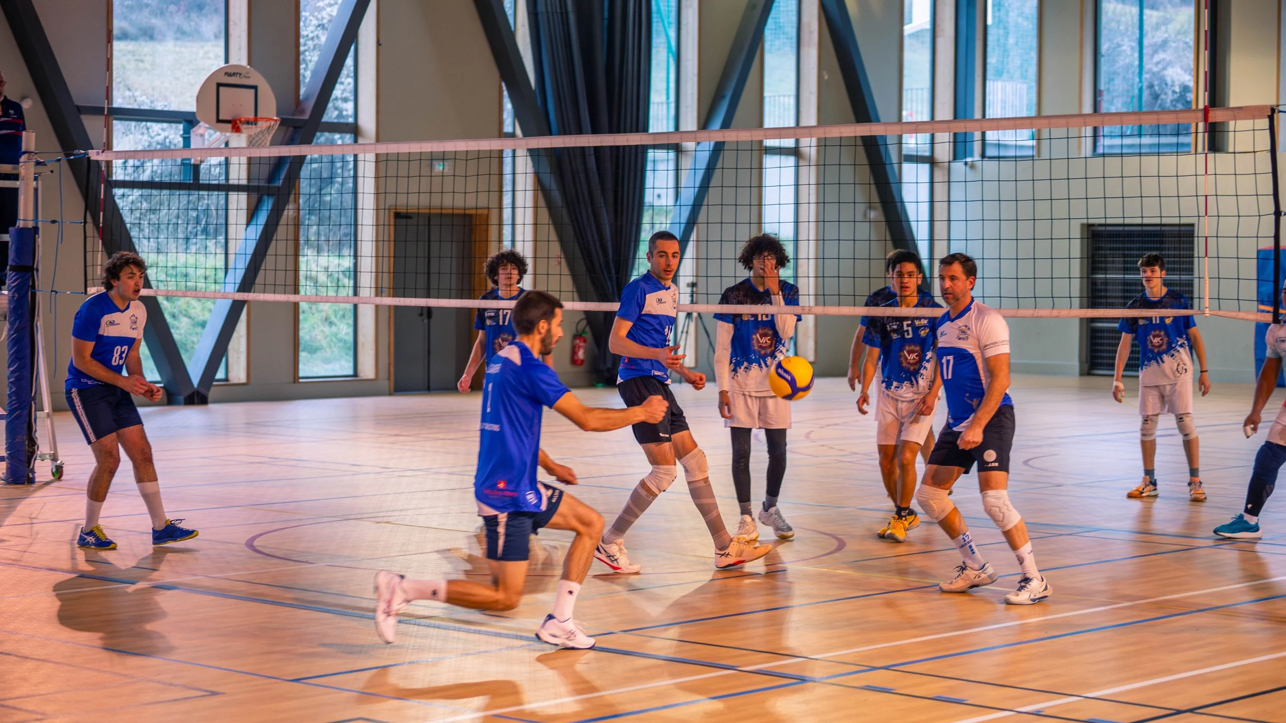 Des jeunes joueurs de volleyball en action dans un gymnase, portant des maillots bleus et blancs, avec un filet de volley-ball entre eux.