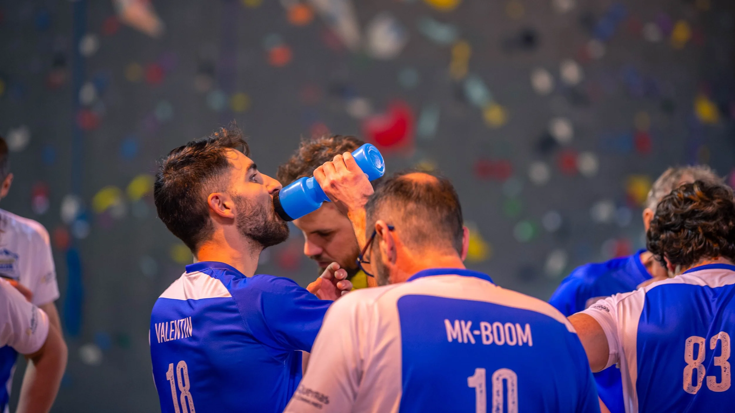 Une équipe de volleyball en tenues bleues, certains membres buvant de l'eau pendant une pause dans le match, avec un mur d'escalade en arrière-plan.