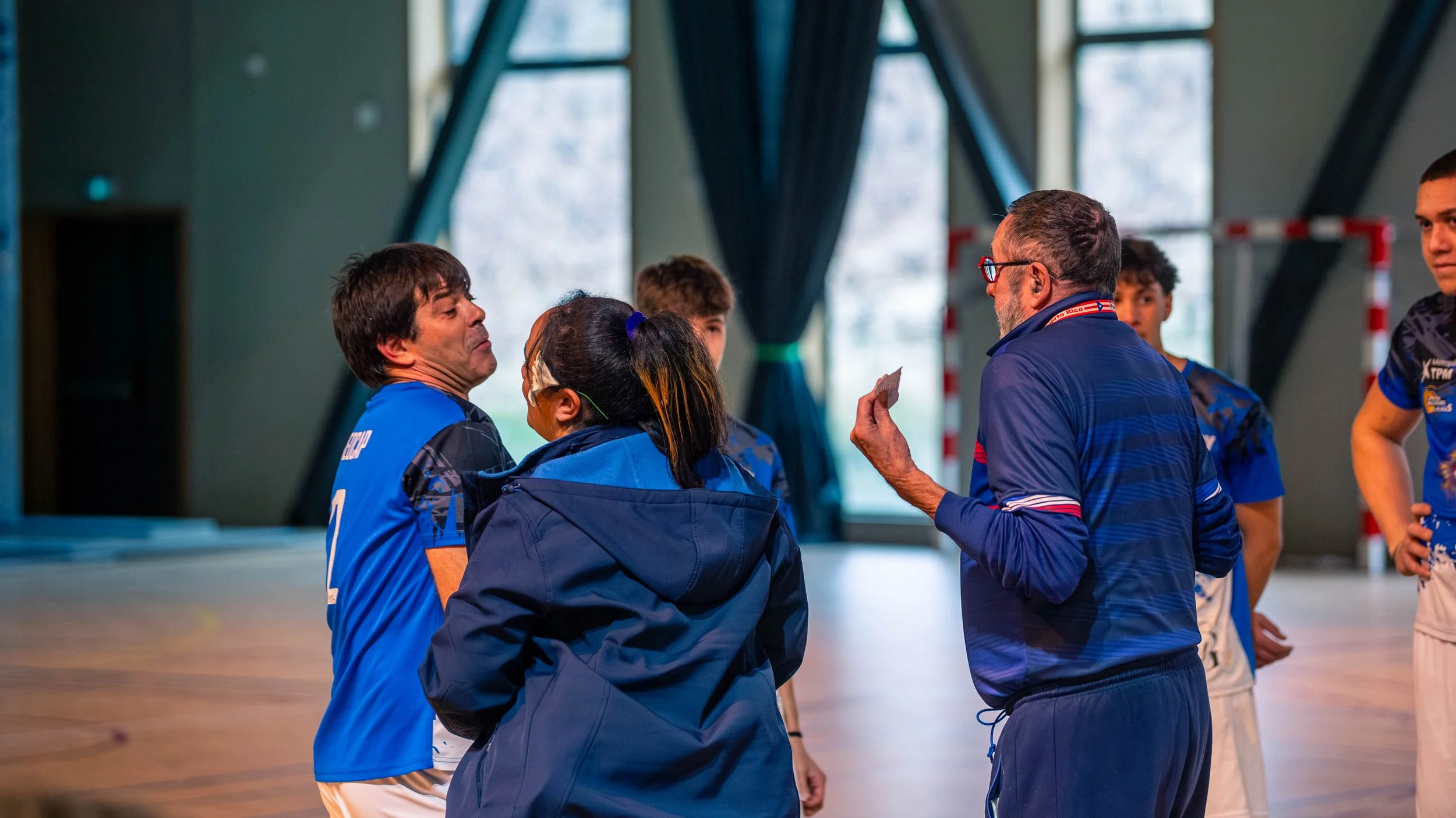 Un entraîneur parle à une équipe de joueurs de handball dans une salle intérieure, probablement lors d'une pause ou d'une stratégie, avec des fenêtres en arrière-plan et un but de handball.