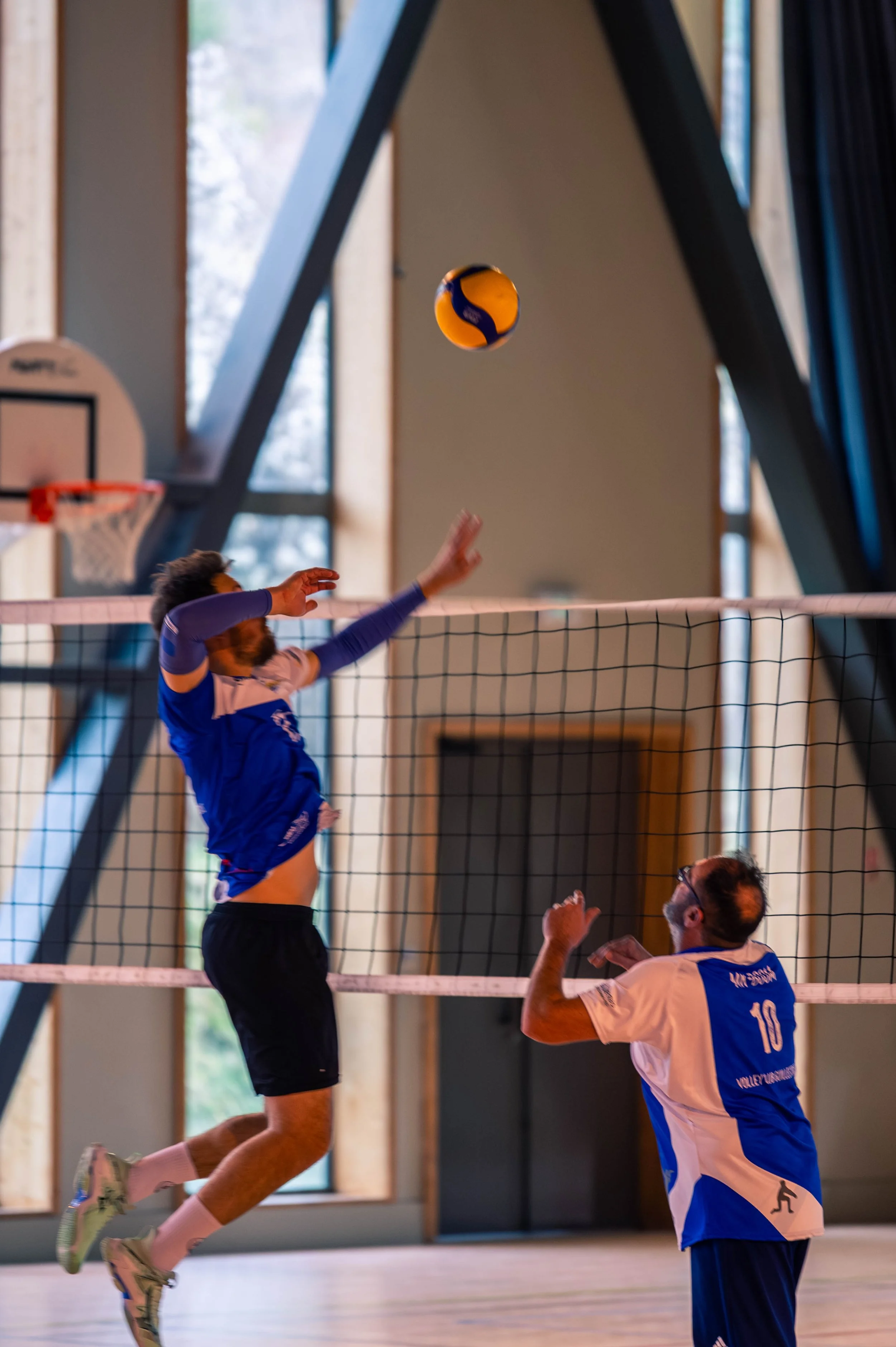 Deux joueurs de volleyball en action dans une salle, un d'eux saute pour frapper le ballon, l'autre se prépare à recevoir.