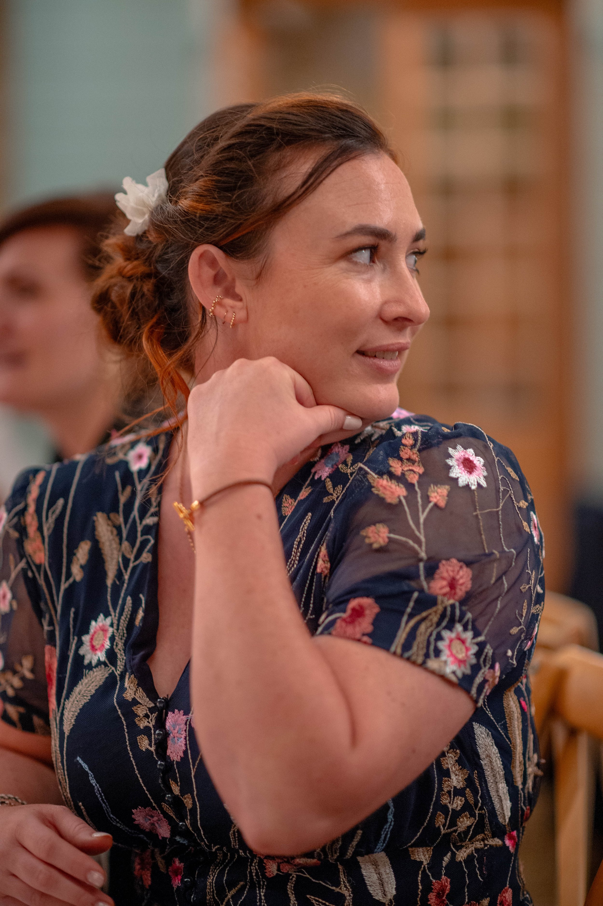 Femme souriante à côté d'une autre femme dans une salle, portant une robe à fleurs et une coiffure tressée avec un ruban blanc.