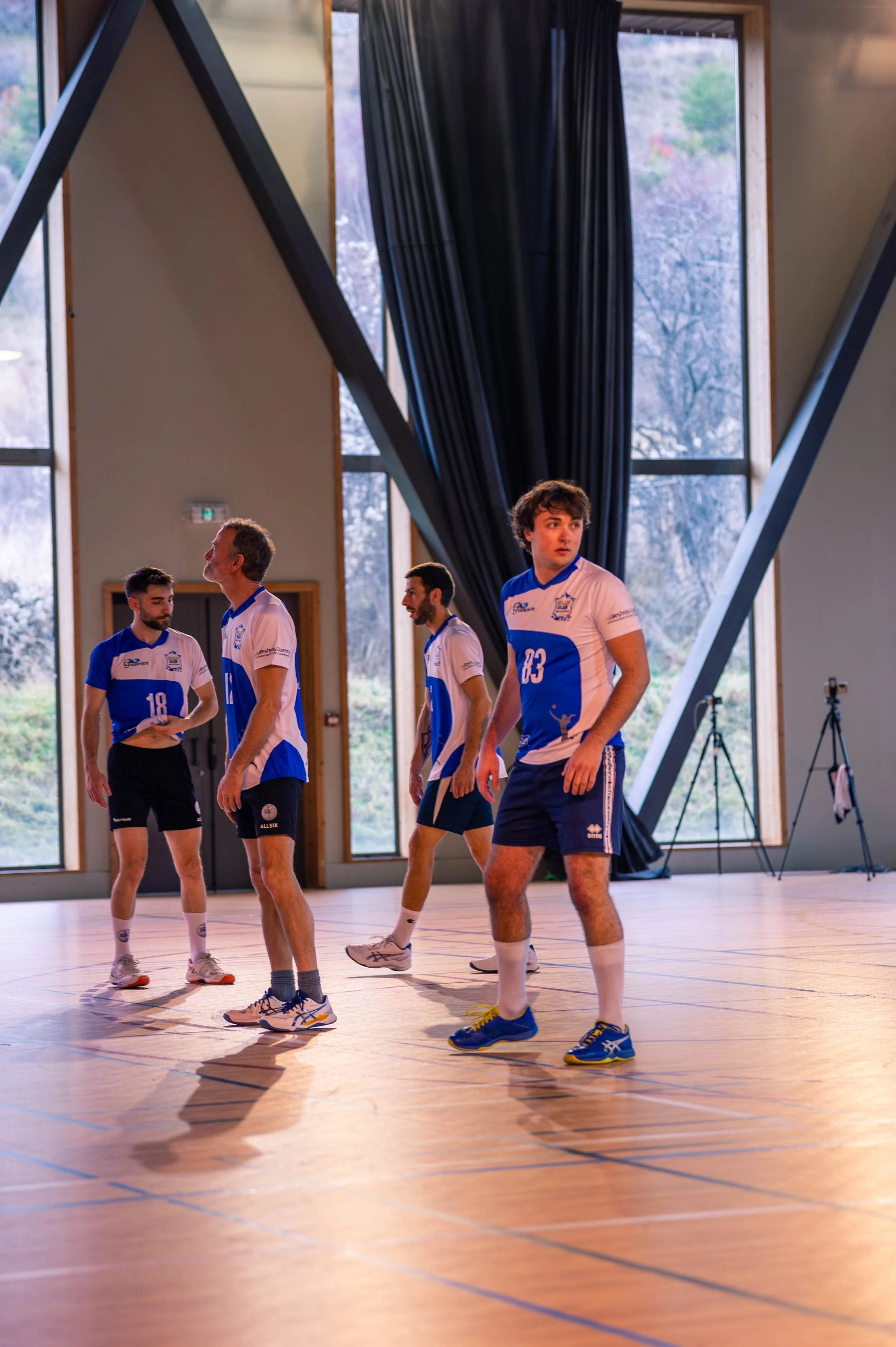 Jeu de volleyball en intérieur avec quatre joueurs en tenue sportive bleue et blanche, dans une salle avec de grandes fenêtres et rideaux noirs.