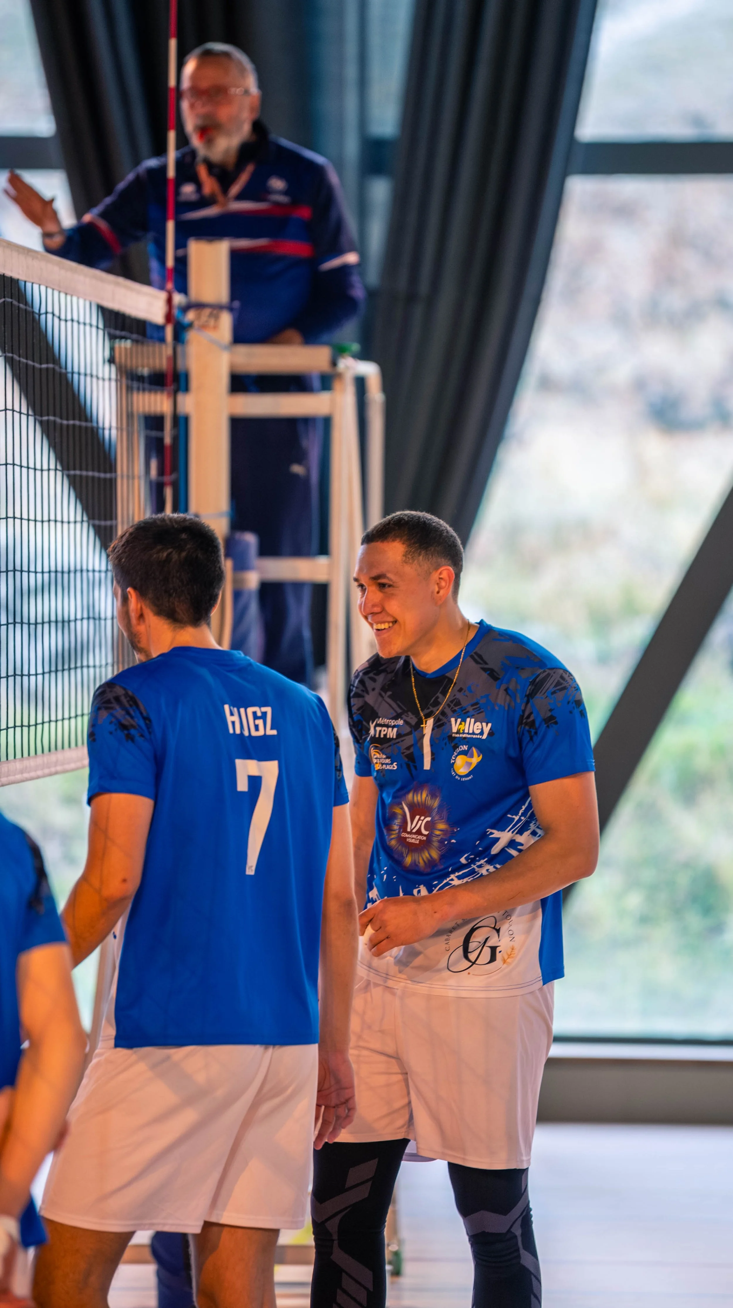 Deux joueurs de volleyball en uniforme bleu, souriants, discutent près d'un filet lors d'un match ou d'un entraînement, avec un entraîneur en arrière-plan, dans une salle avec de grandes fenêtres en arrière-plan.