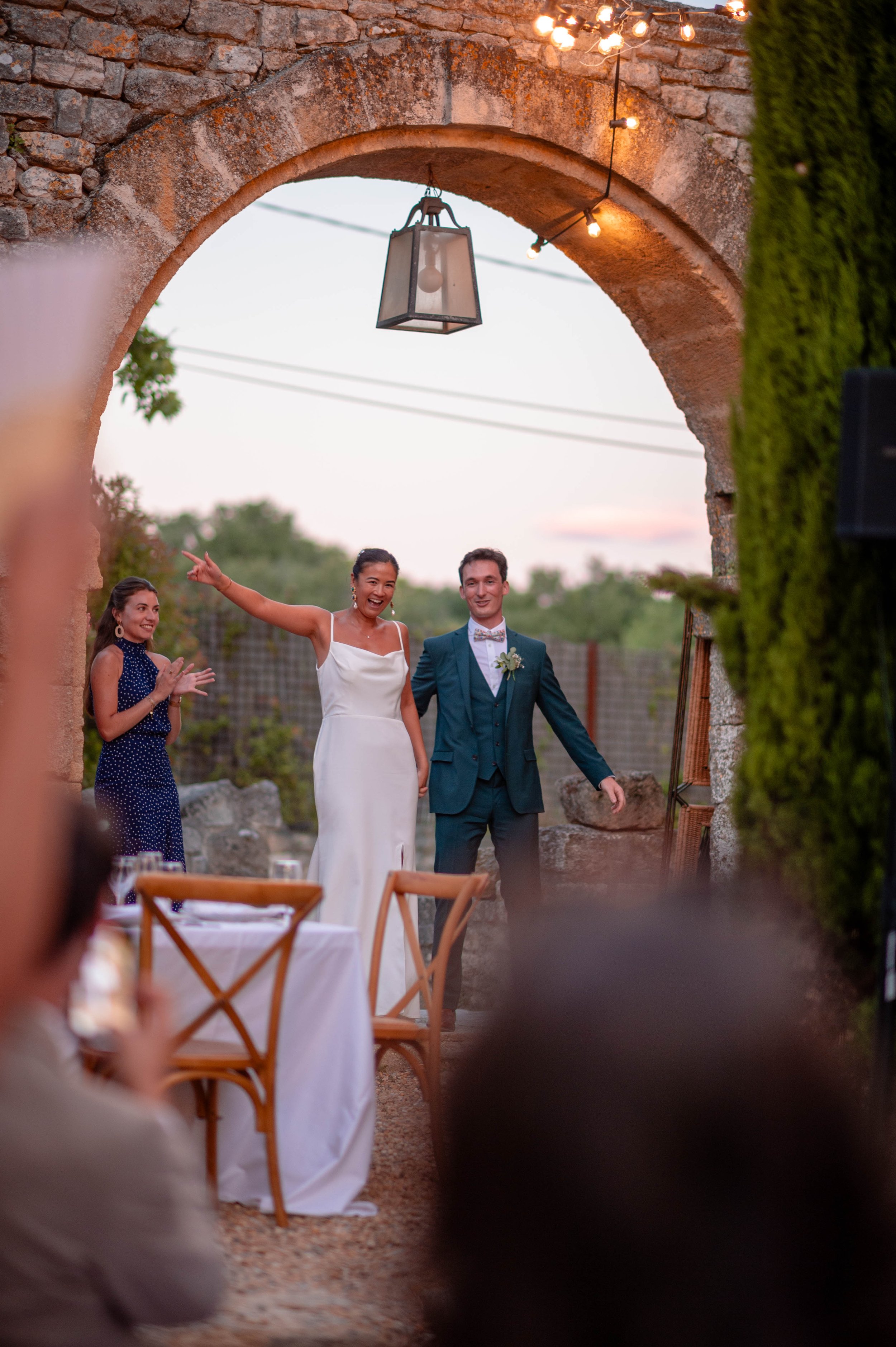 Un couple de mariés souriant lors de leur mariage en extérieur, entouré de deux femmes applaudissant, dans un cadre rustique avec une arche en pierre et des guirlandes lumineuses au coucher du soleil.