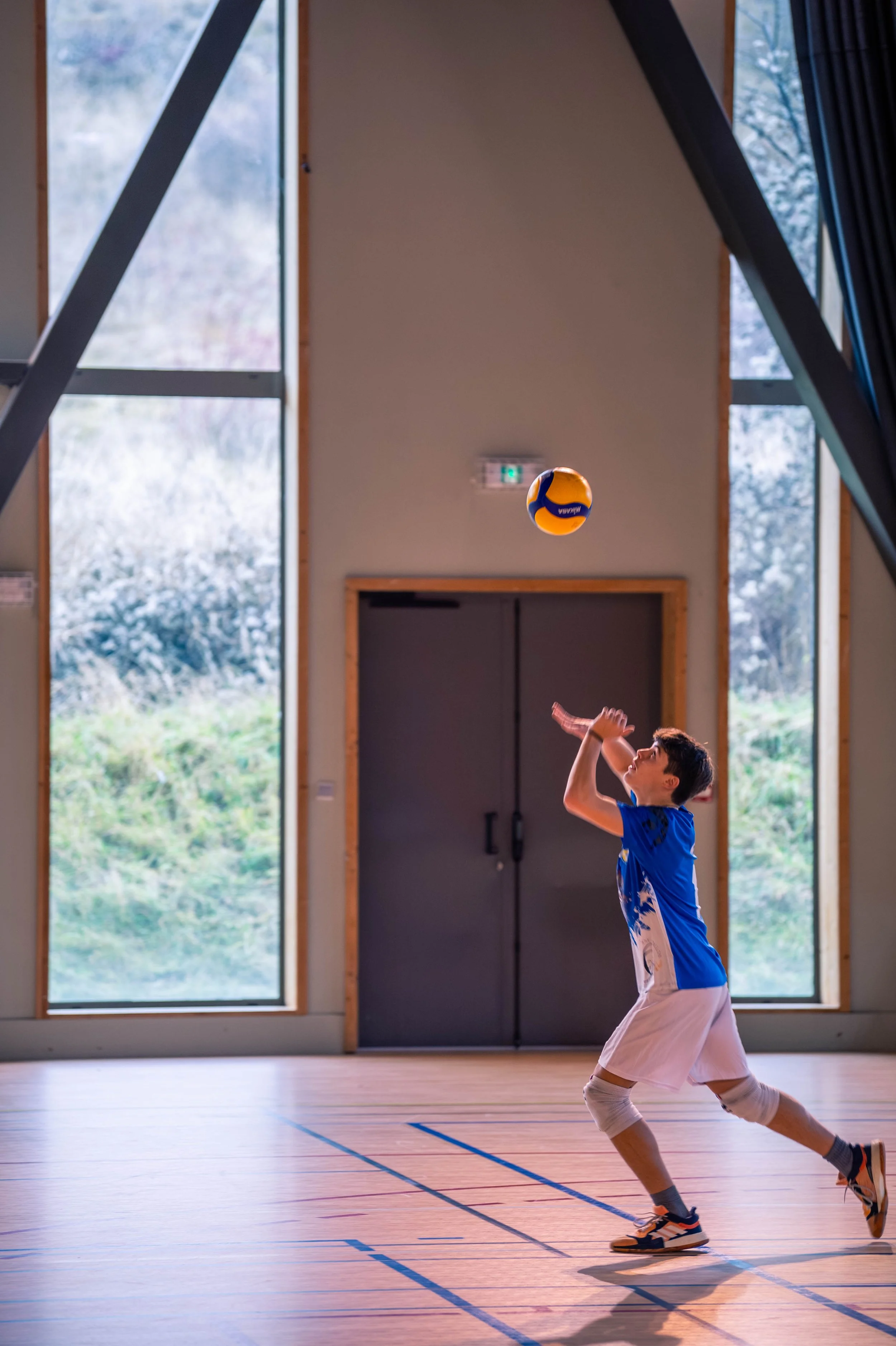 Un jeune joueur en tenue de volley-ball en train de faire un service dans un gymnase intérieur. Il porte un maillot bleu, un short blanc et des genouillères, avec un ballon de volleyball orange et bleu en suspension dans l'air.
