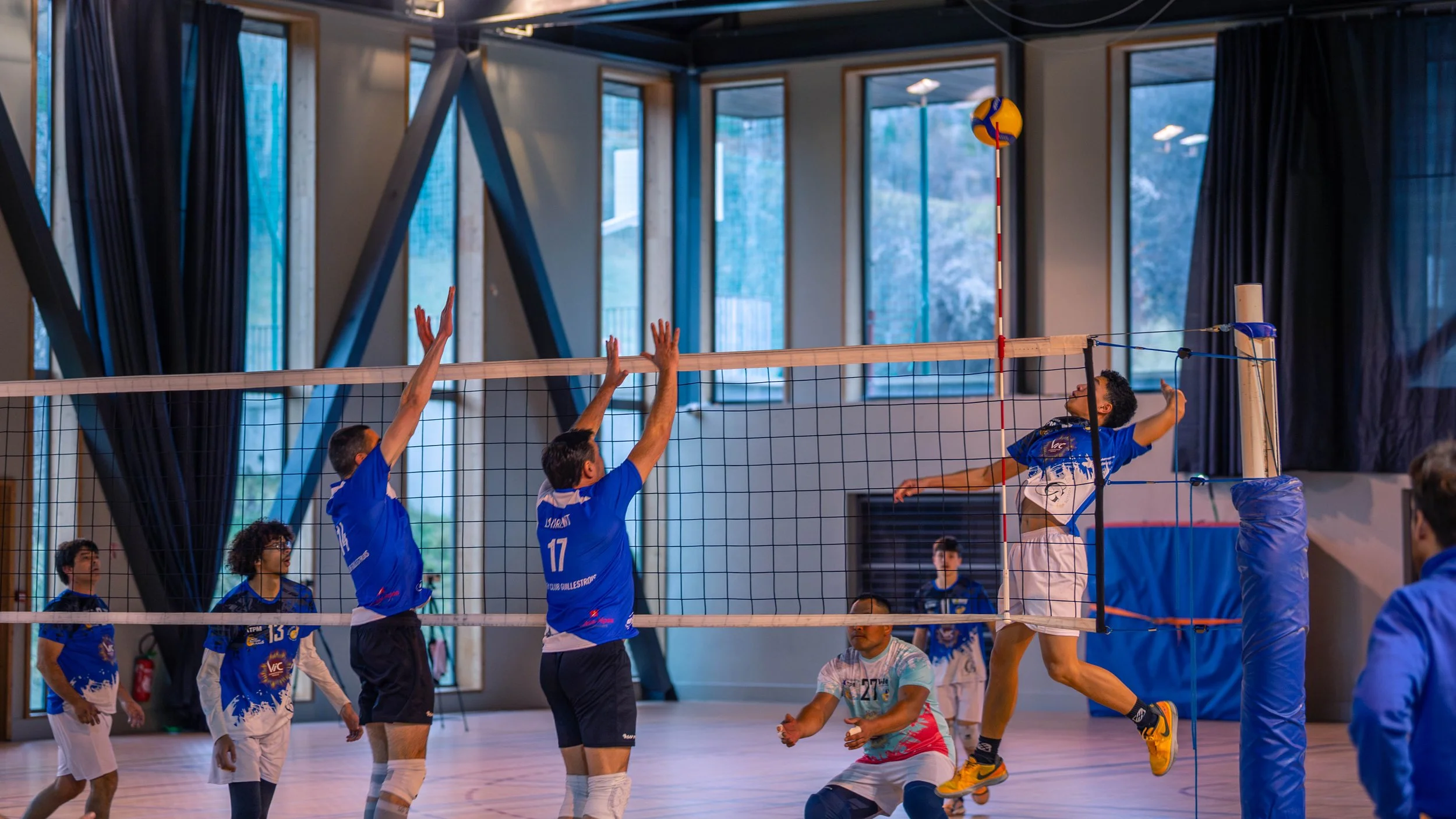 Des joueurs de volleyball en action durant un match en intérieur, avec un joueur en saut tentant de frapper le ballon, d'autres joueurs prêts à bloquer ou à recevoir, dans une salle avec grandes fenêtres et rideaux noirs.