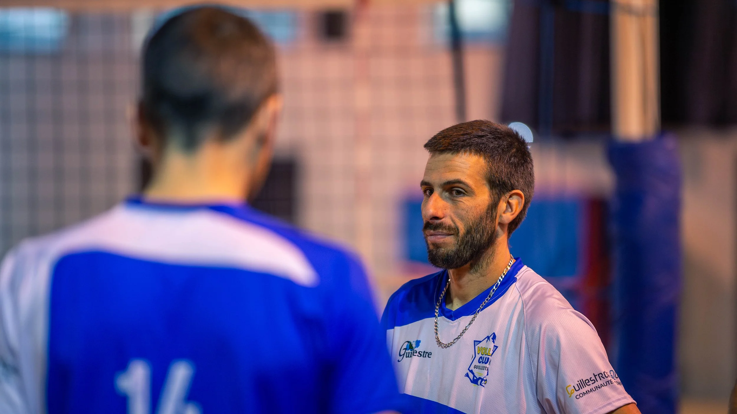 Deux hommes portent des maillots de sport bleus et blancs lors d'une séance d'entraînement ou d'une rencontre sportive en intérieur.