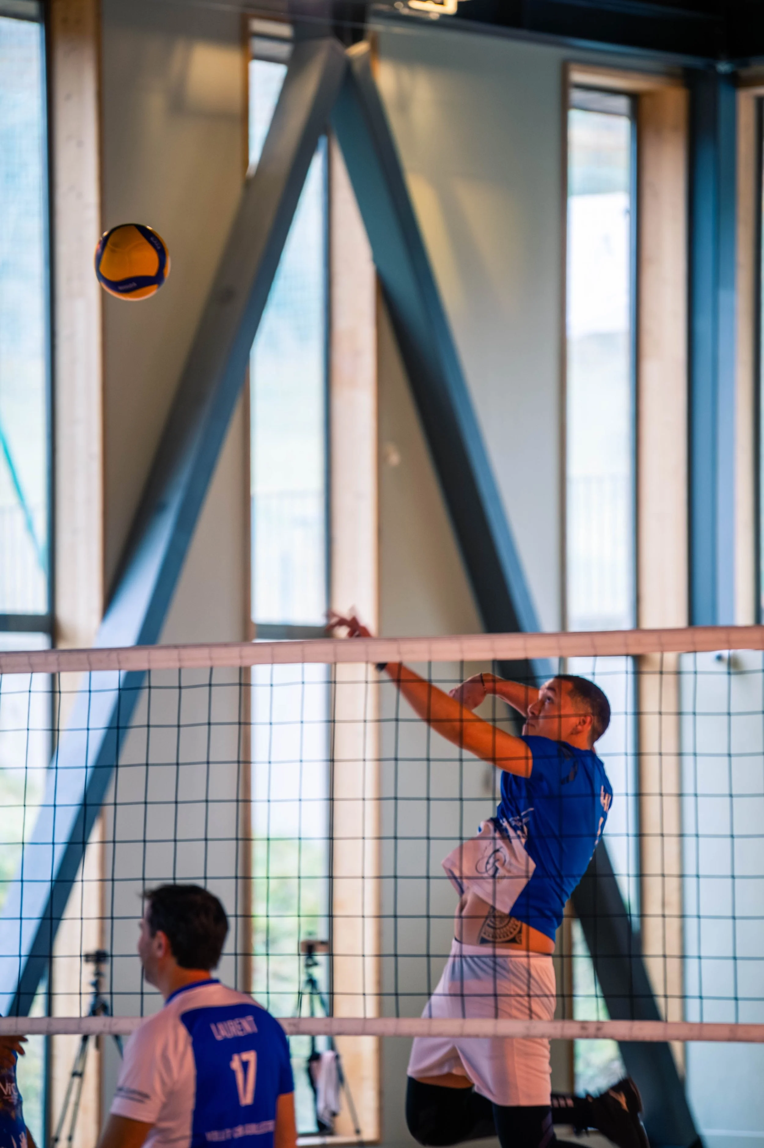 Deux joueurs de volley-ball en train de jouer, un saut pour frapper le ballon au-dessus du filet dans un gymnase, avec des fenêtres en arrière-plan.